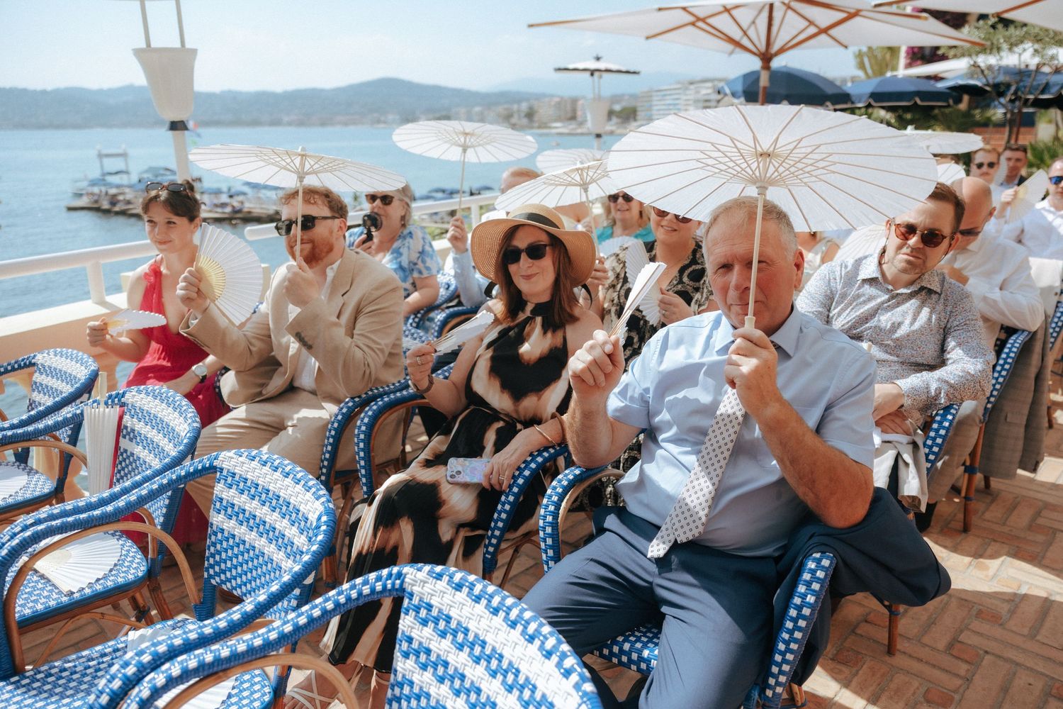 People enjoying drinks at an outdoor coastal restaurant with white umbrellas and blue and white chairs by the sea.