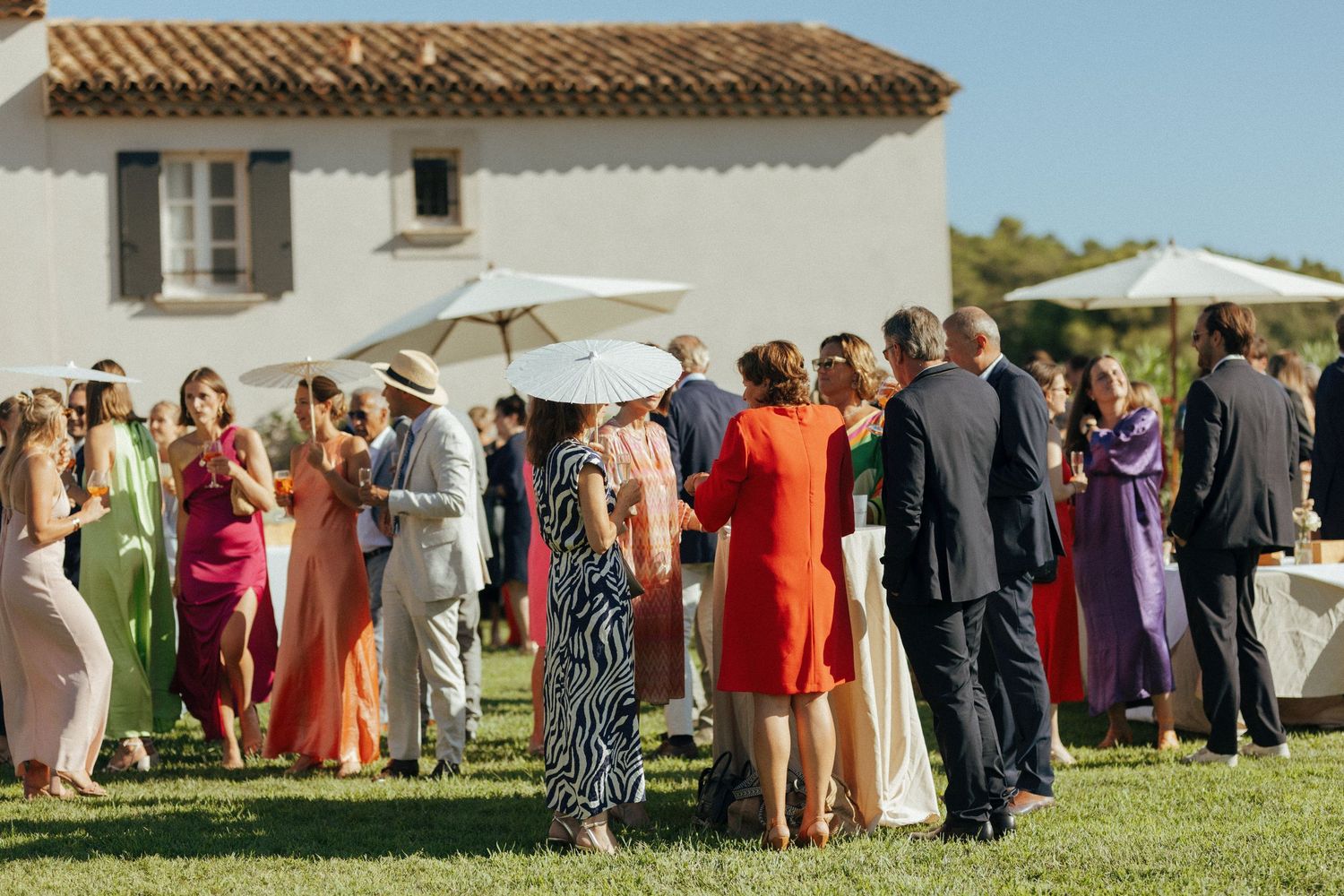 Wedding guests mingle outside a Mediterranean villa with colorful dresses and parasols.