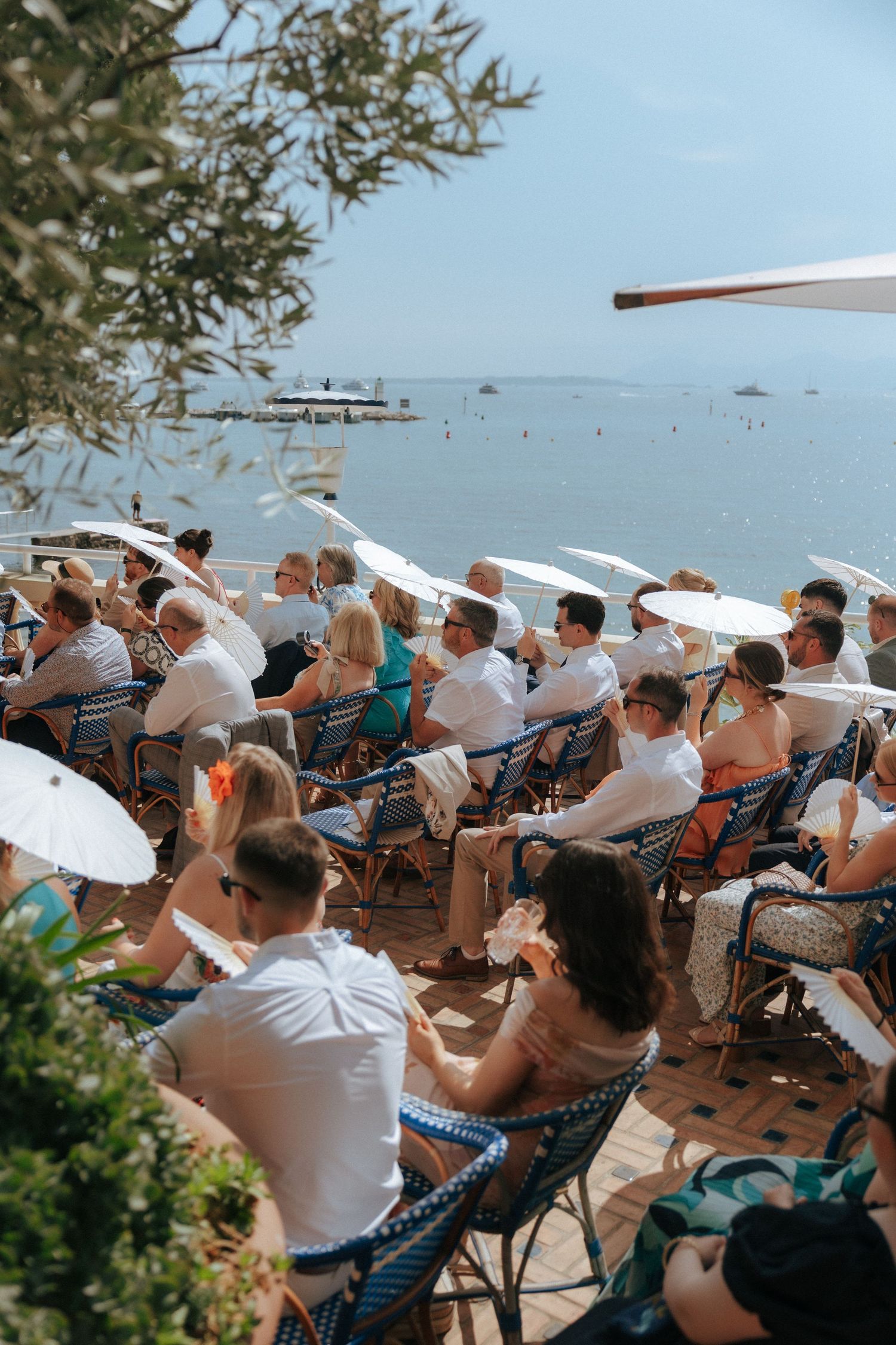Wedding guests holding white parasols while seated at an outdoor seaside ceremony.
