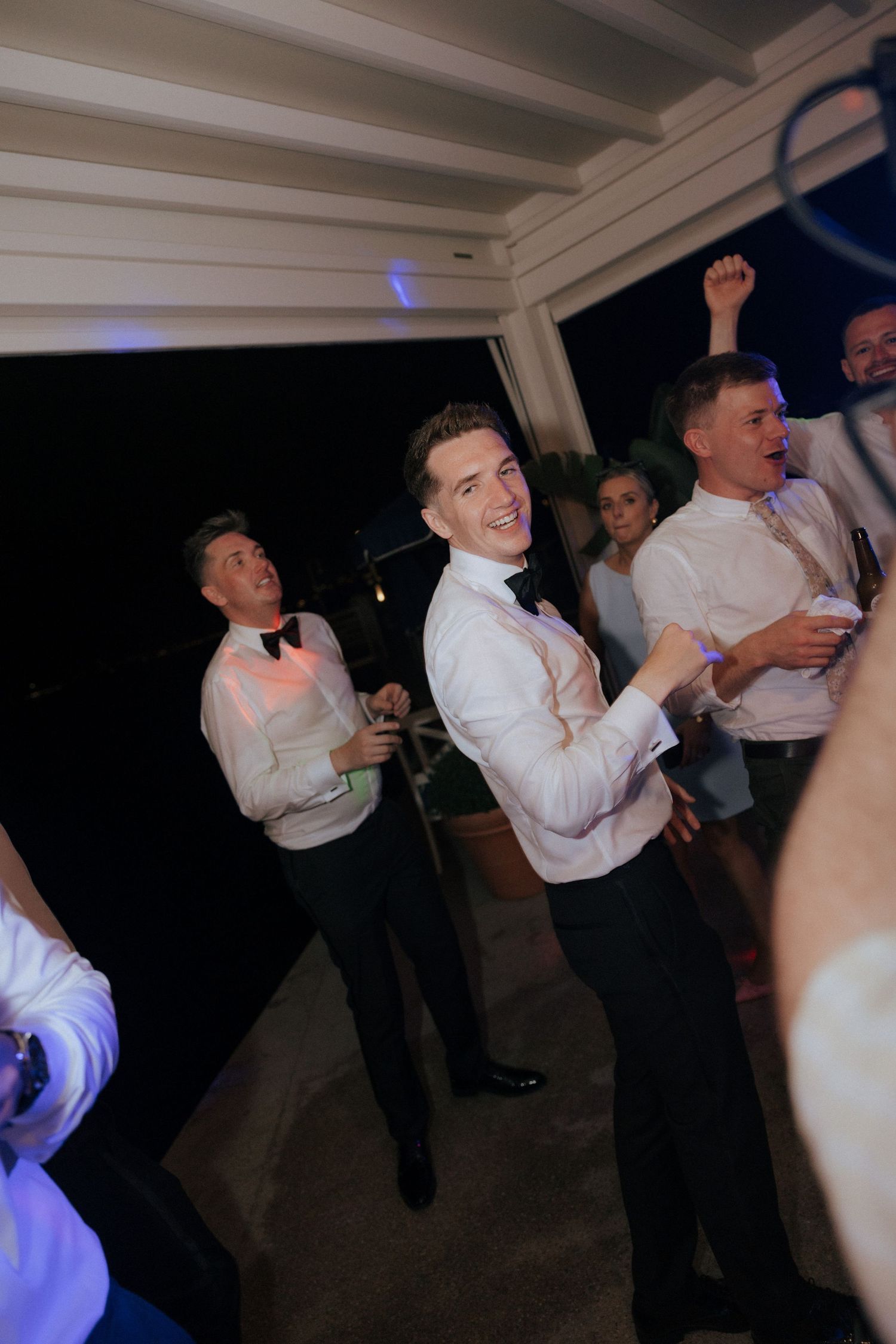 Wedding guests dance energetically at a dimly lit reception party.