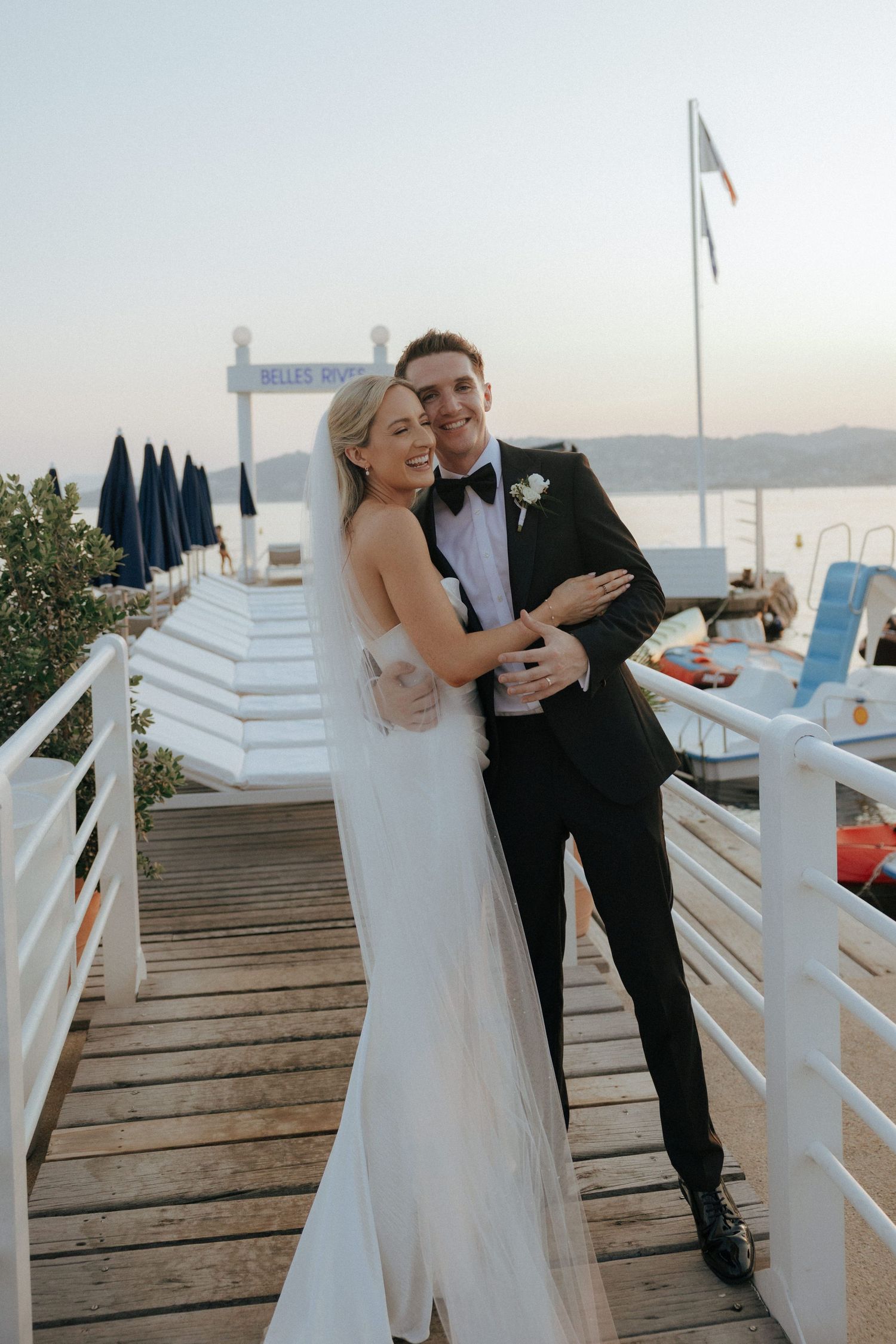 A couple shares a romantic moment on a wooden deck overlooking the water during their wedding celebration.