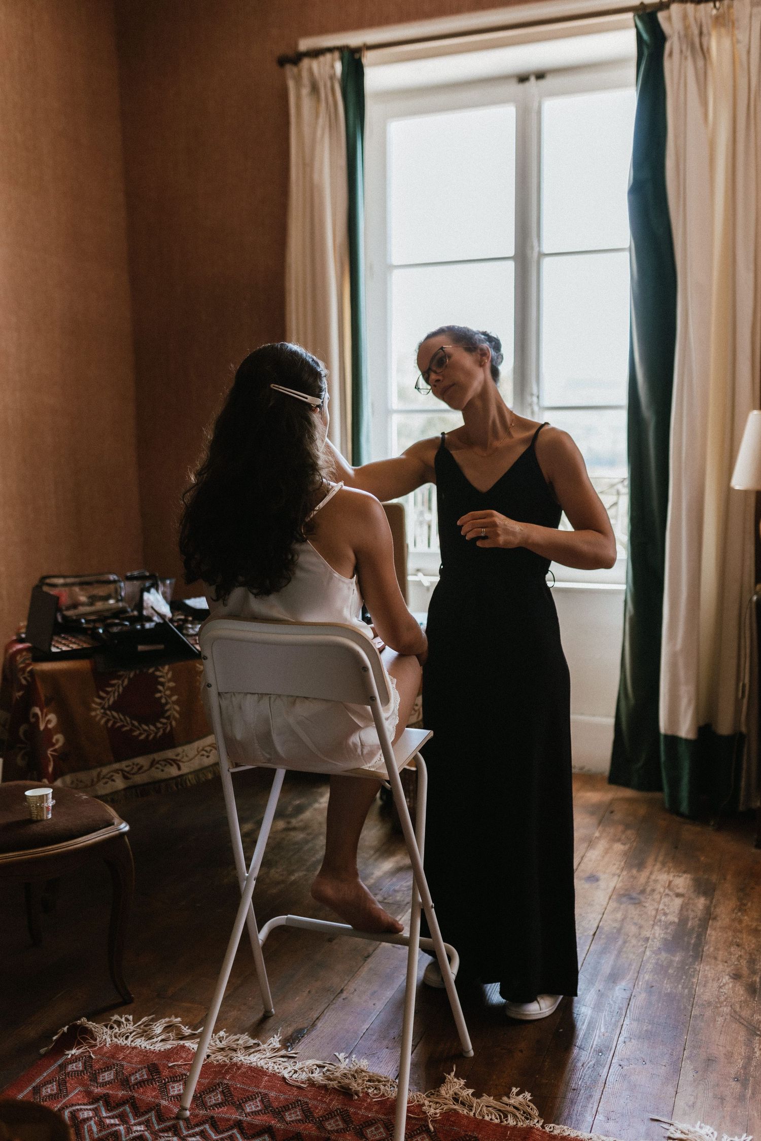 Two people in a warmly lit room preparing for a formal event near a window with teal curtains.