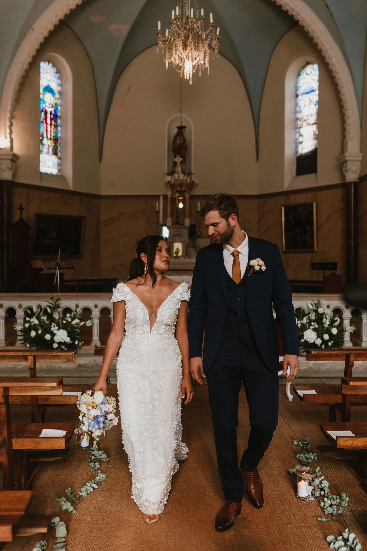 A newly married couple walking down the aisle in a chapel with stained glass windows and floral decor.