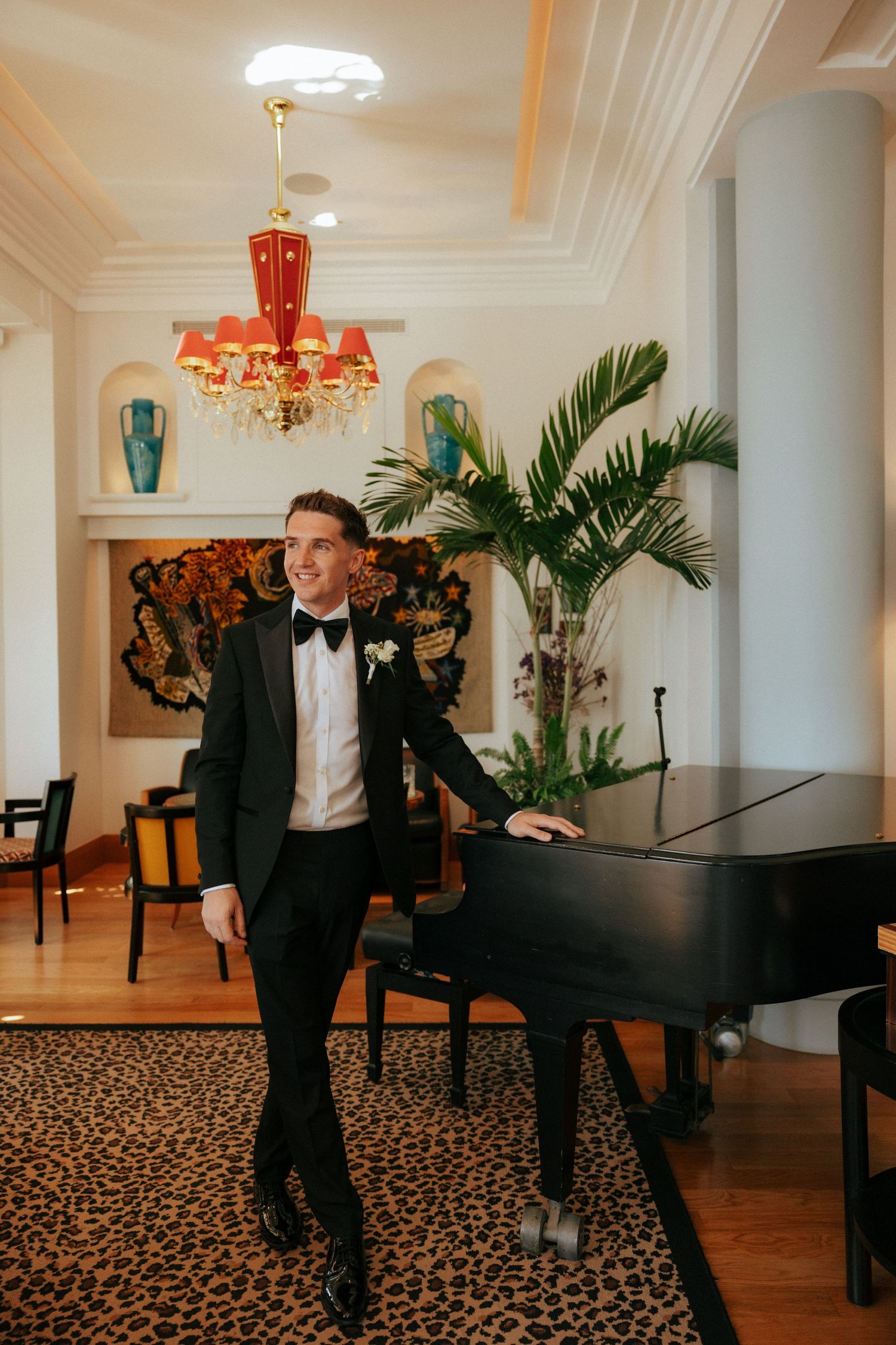 A groom in formal attire poses playfully in an elegant room with palm plants and a grand piano.