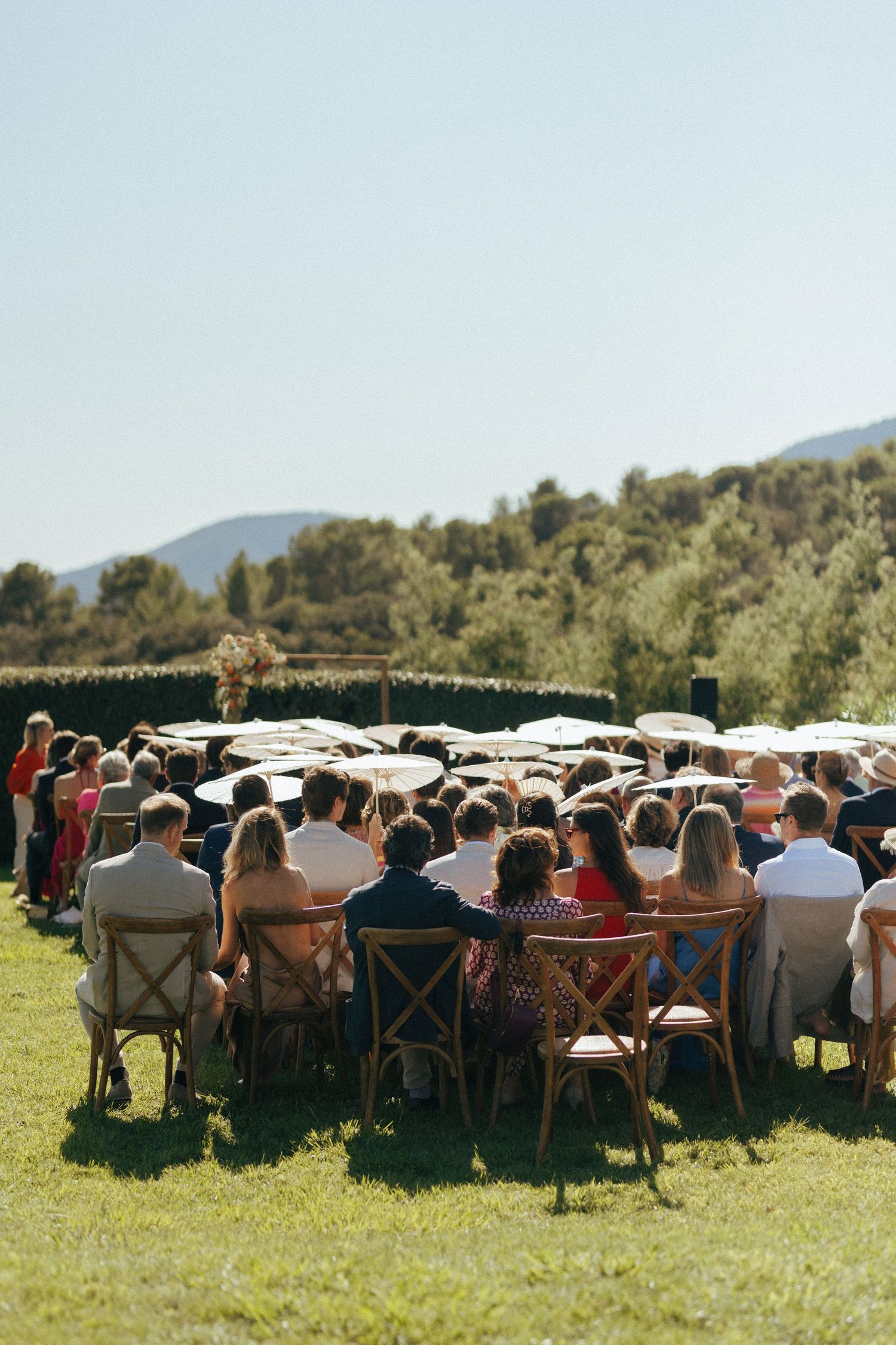 Wedding guests seated in wooden chairs on a grassy lawn for an outdoor ceremony with mountains in the background.