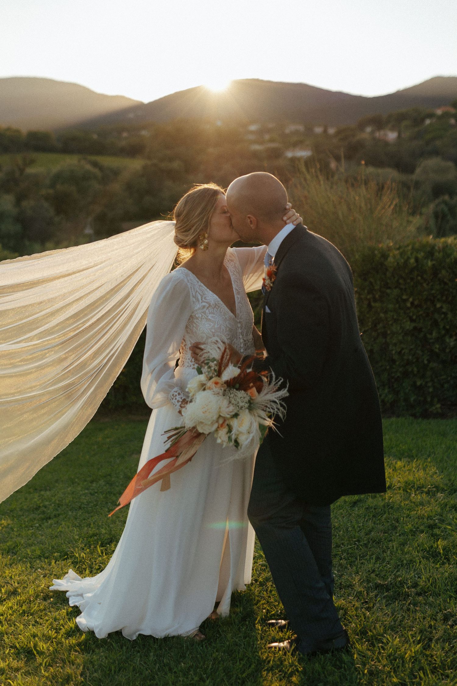 Romantic sunset wedding photo with flowing veil and mountain backdrop.