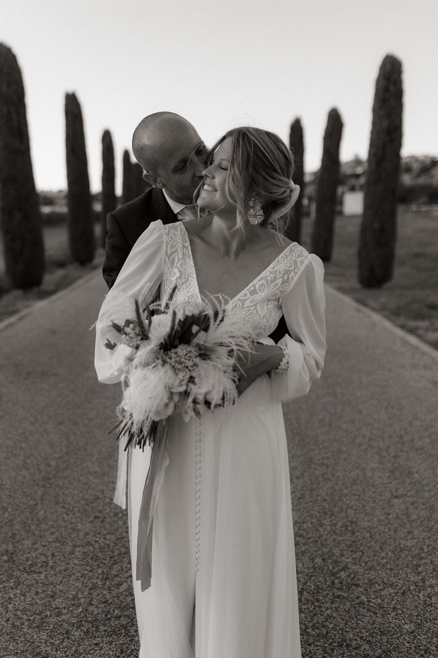 Romantic black and white wedding portrait with cypress trees lining pathway.