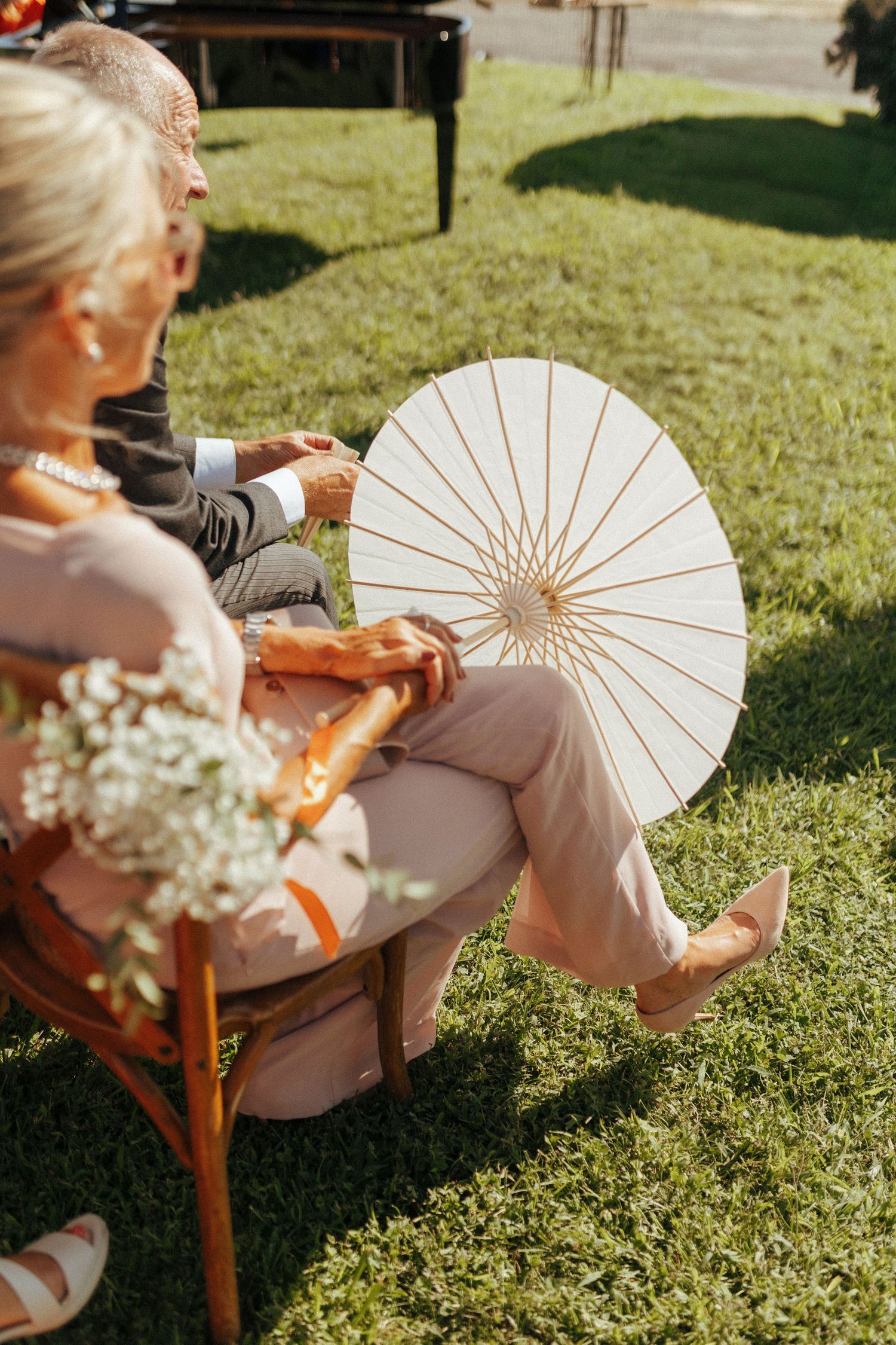 A white paper parasol is held on a sunny lawn while guests sit in wooden chairs at an outdoor gathering.