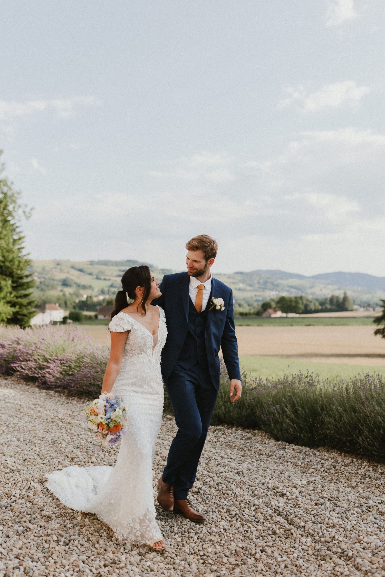 A couple walks along a gravel path with rolling hills in the background on their wedding day.