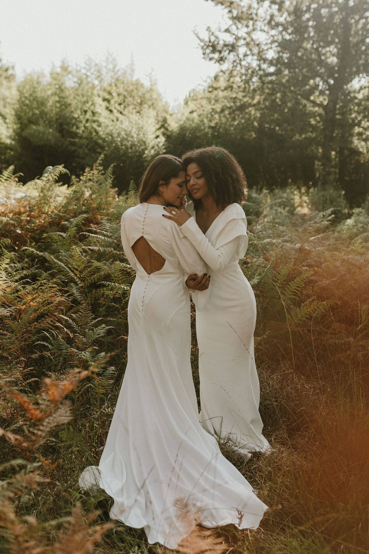 Two brides in flowing white wedding gowns share an intimate moment among wild ferns at golden hour.