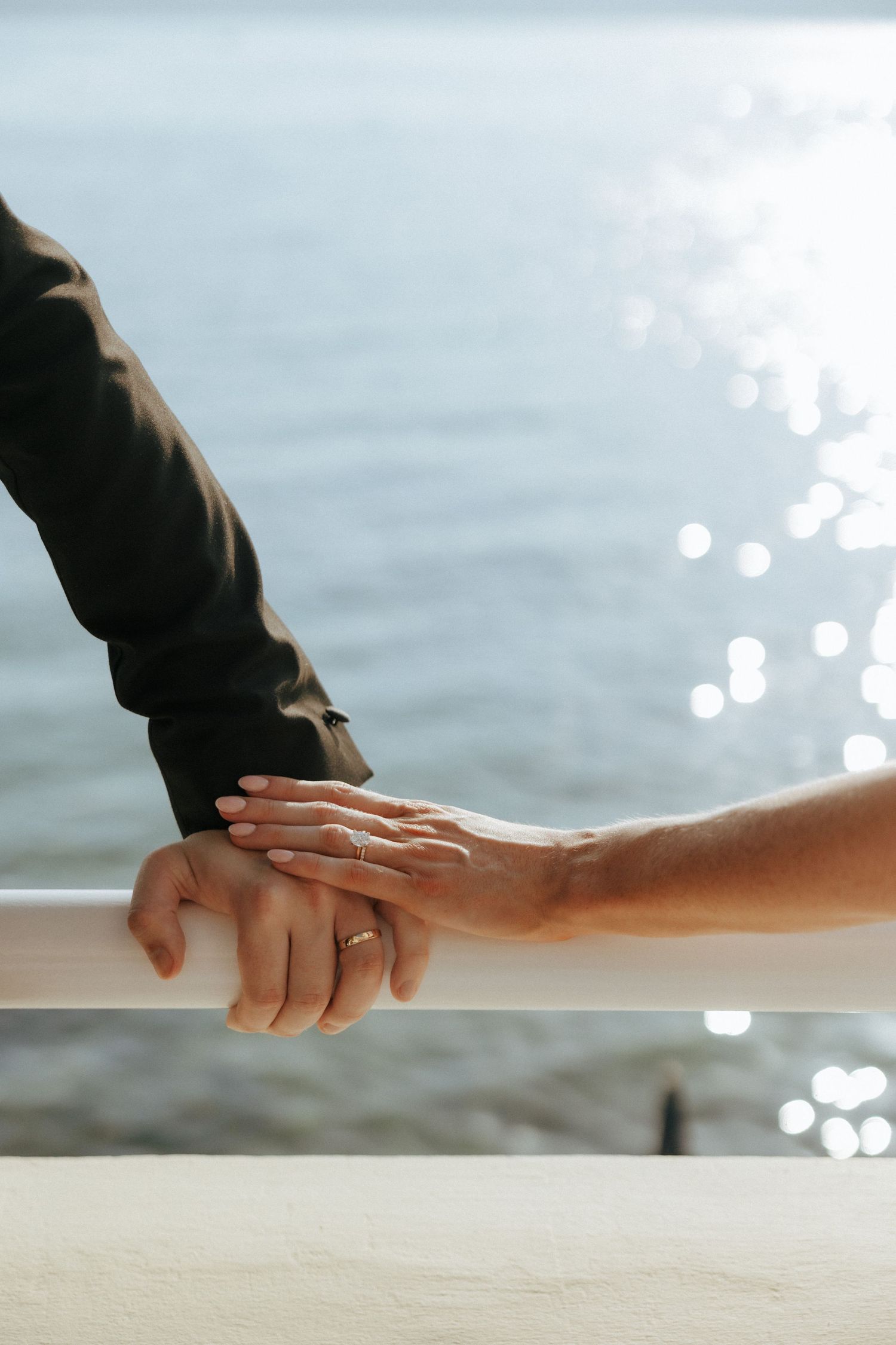 Two hands touch tenderly on a railing with sparkling ocean waters in the background on a sunny day.