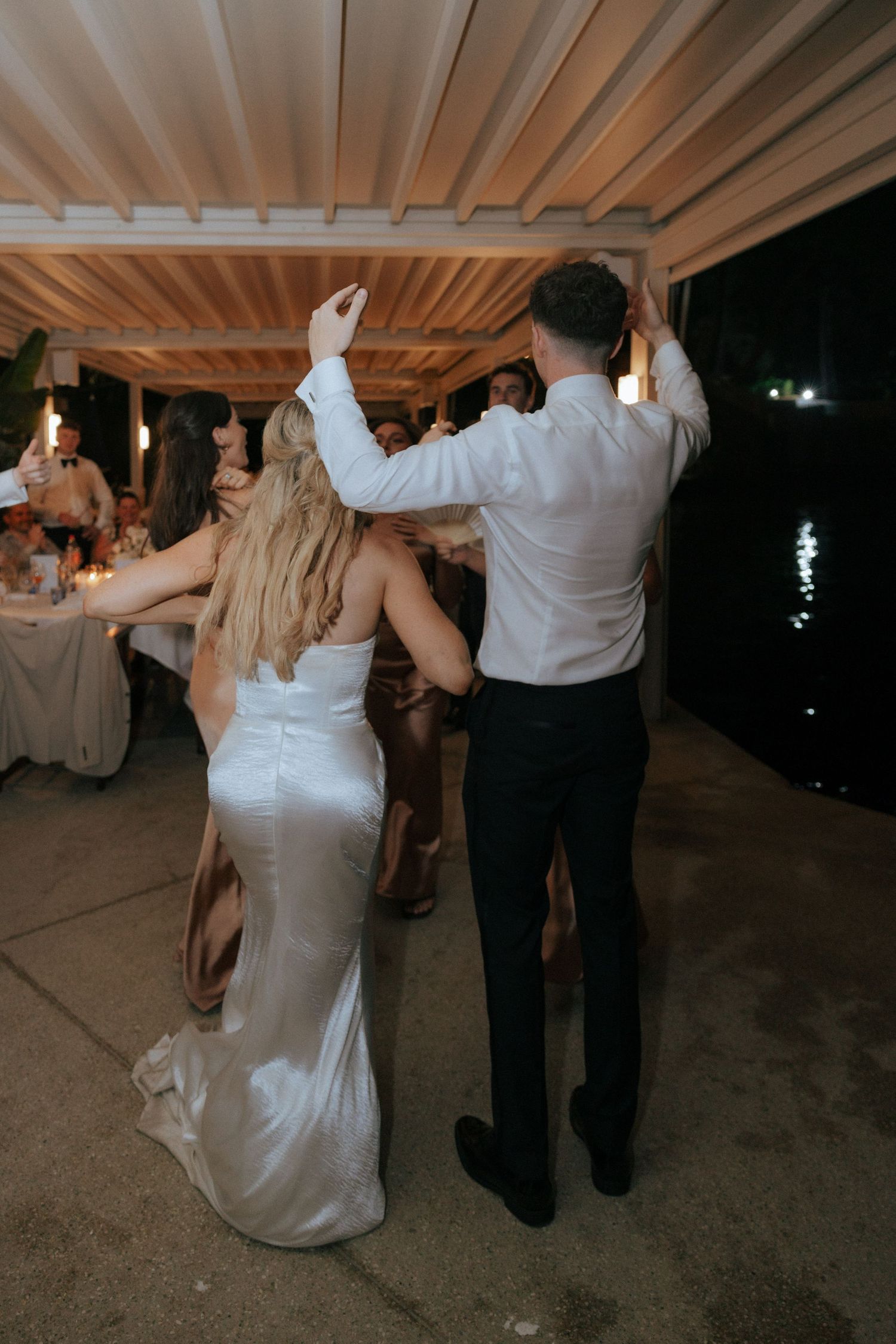 Wedding guests dancing and celebrating at an evening reception under a covered outdoor venue.