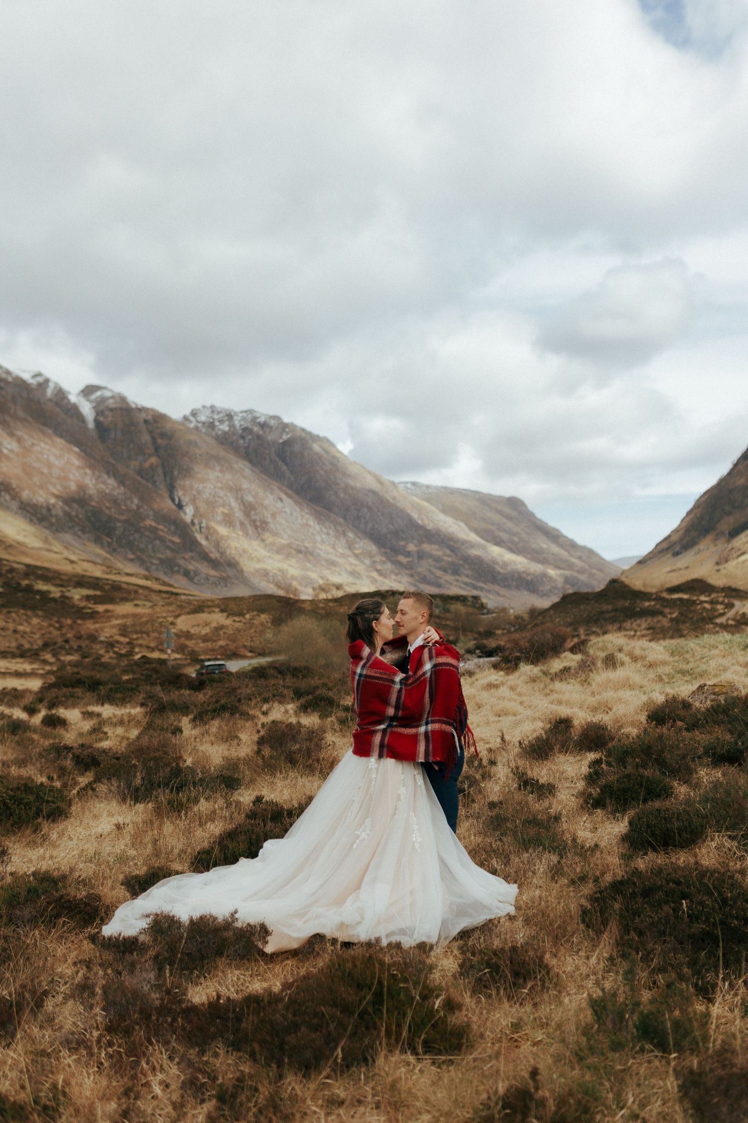 A couple in a traditional red tartan wrap embrace against the dramatic backdrop of the Scottish Highlands on their wedding day.