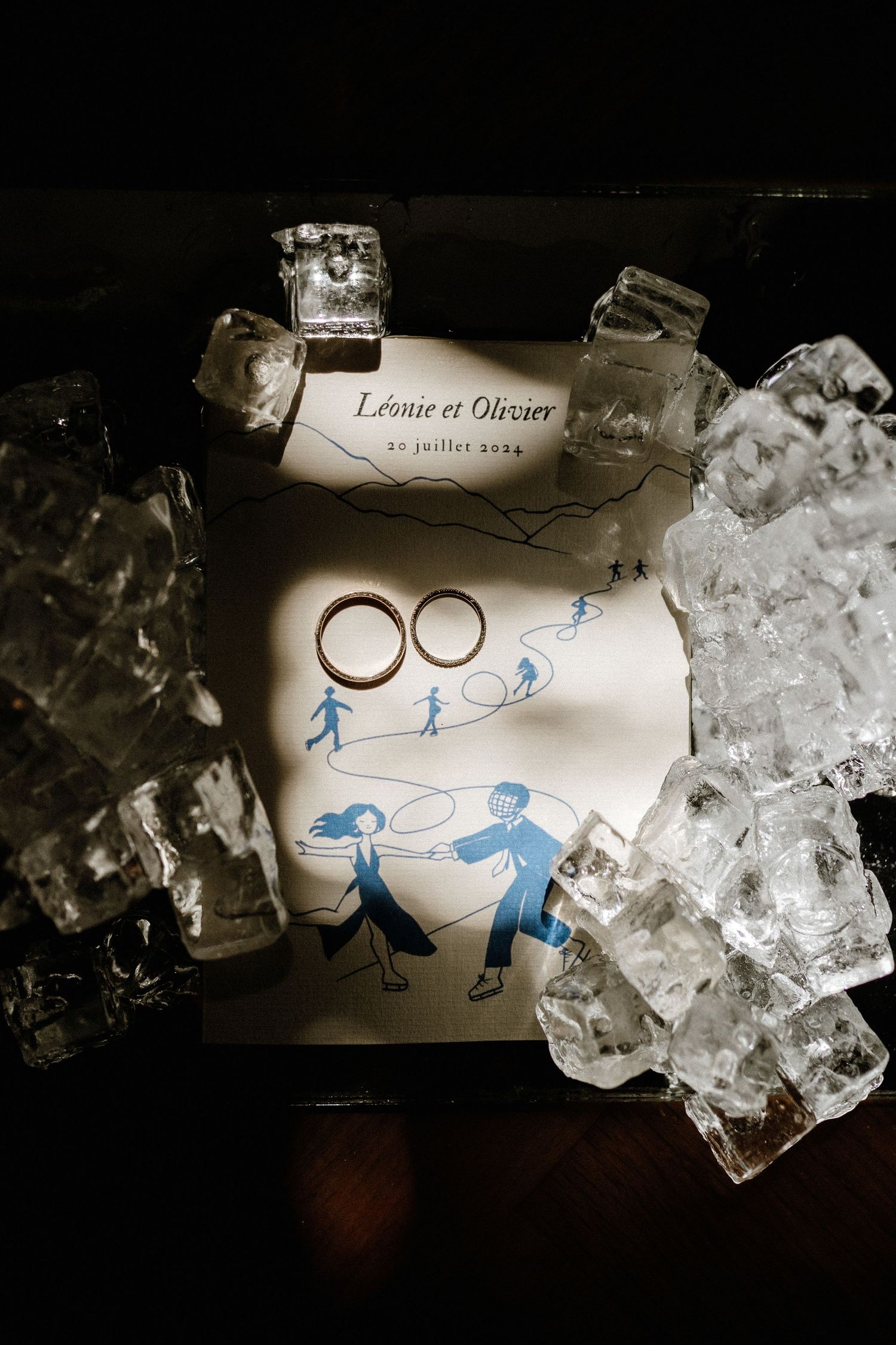Wedding rings and blue stick figure drawings lay surrounded by ice cubes in a dark moody composition.