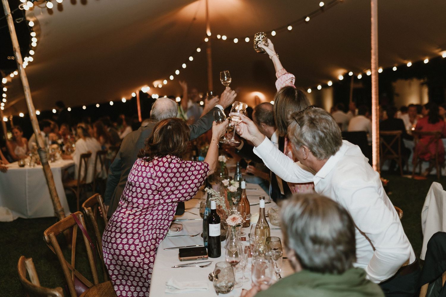 Wedding celebration with guests toasting and raising glasses under a tent with twinkle lights.