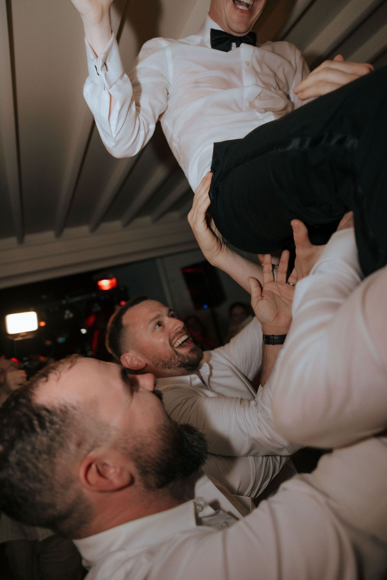 Wedding guests lift someone into the air during an energetic dance celebration indoors.