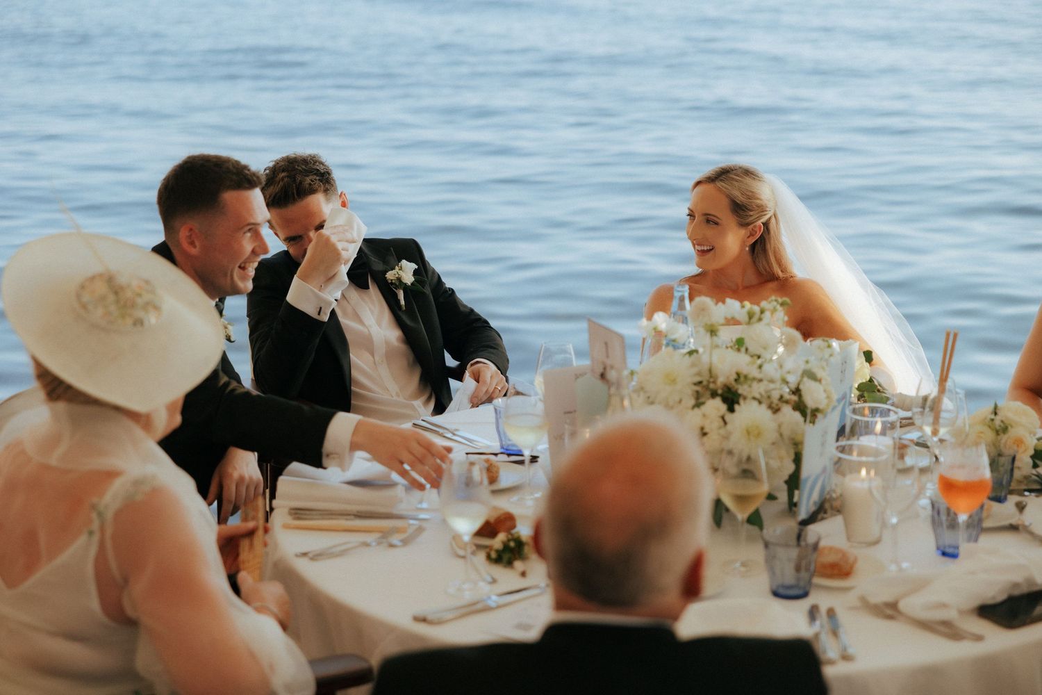 Wedding party gathered at an elegant outdoor reception table by the ocean with white floral centerpieces.