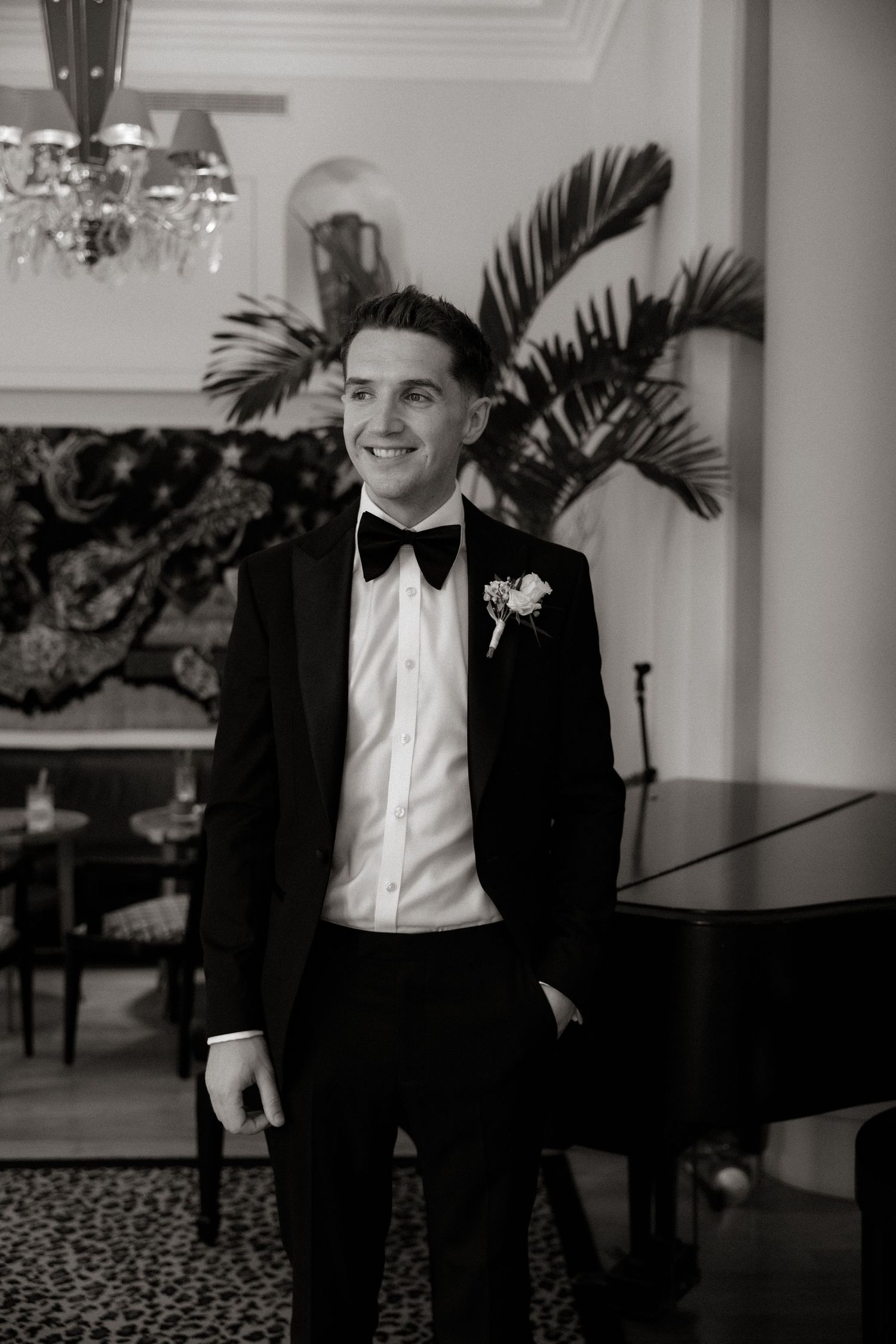 A groom in a classic black tuxedo and bow tie smiles elegantly at a wedding reception with palm decor and piano in background.