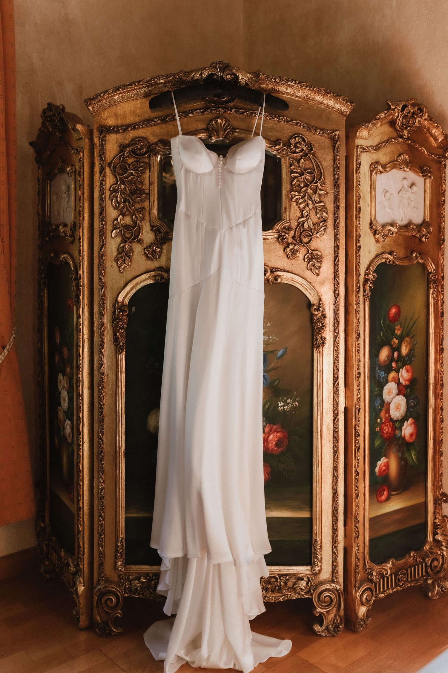 A flowing white wedding dress hangs elegantly in front of an ornate vintage gold mirror with floral painted panels.