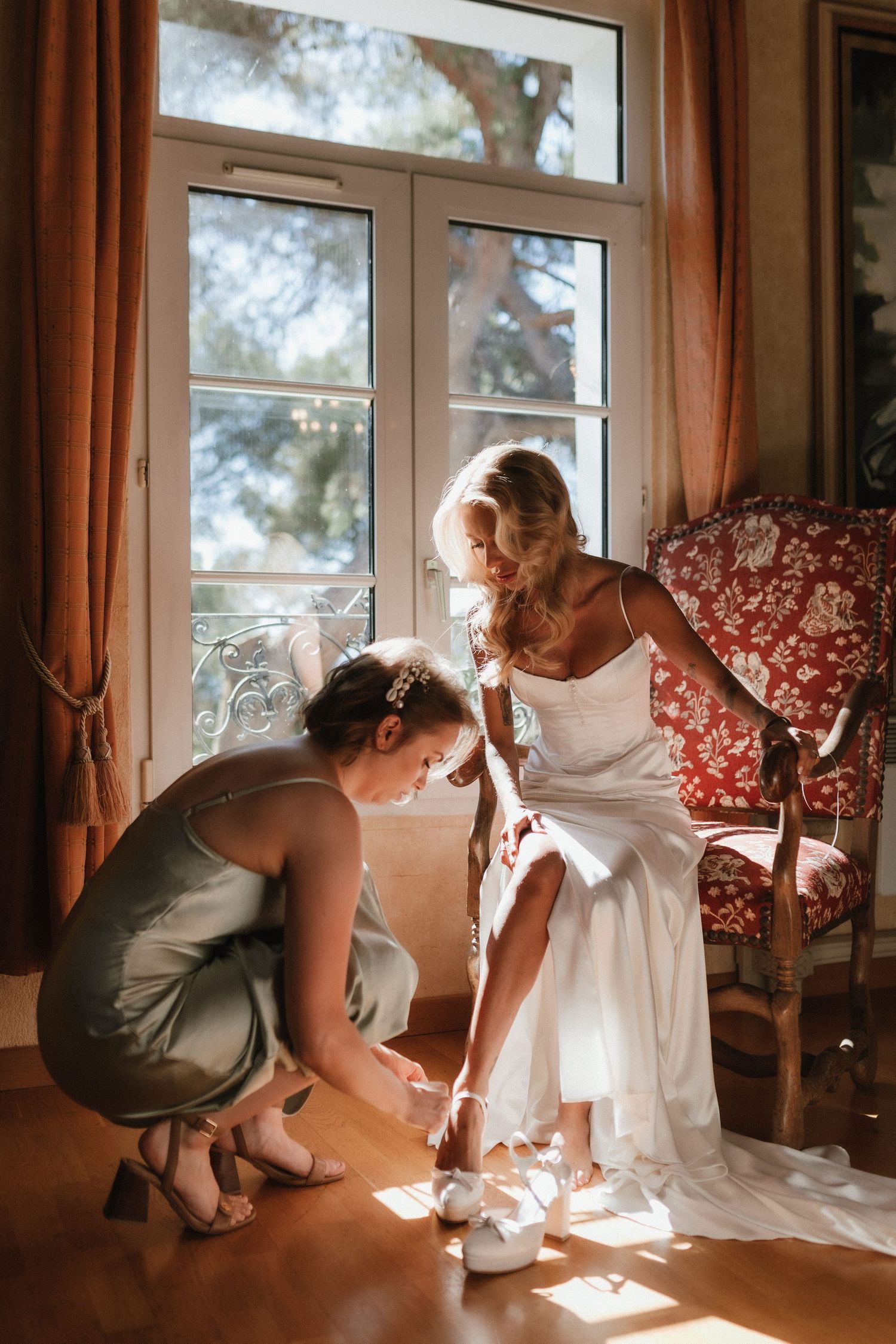 A tender moment in warm sunlight as someone helps adjust shoes in an elegant white dress by a large window.