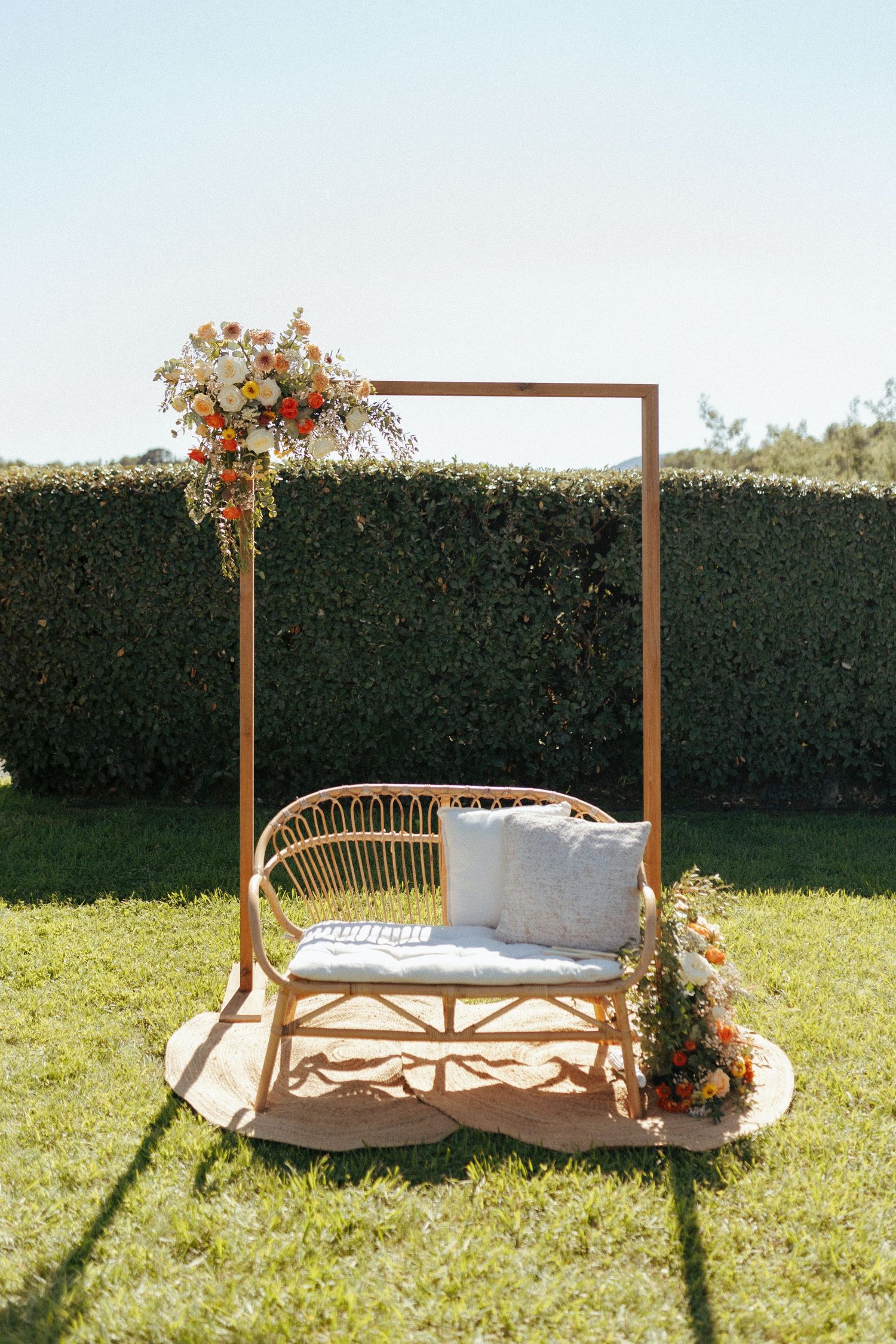 A rustic outdoor wedding ceremony setup with a wooden arch frame and rattan bench decorated with floral arrangements.