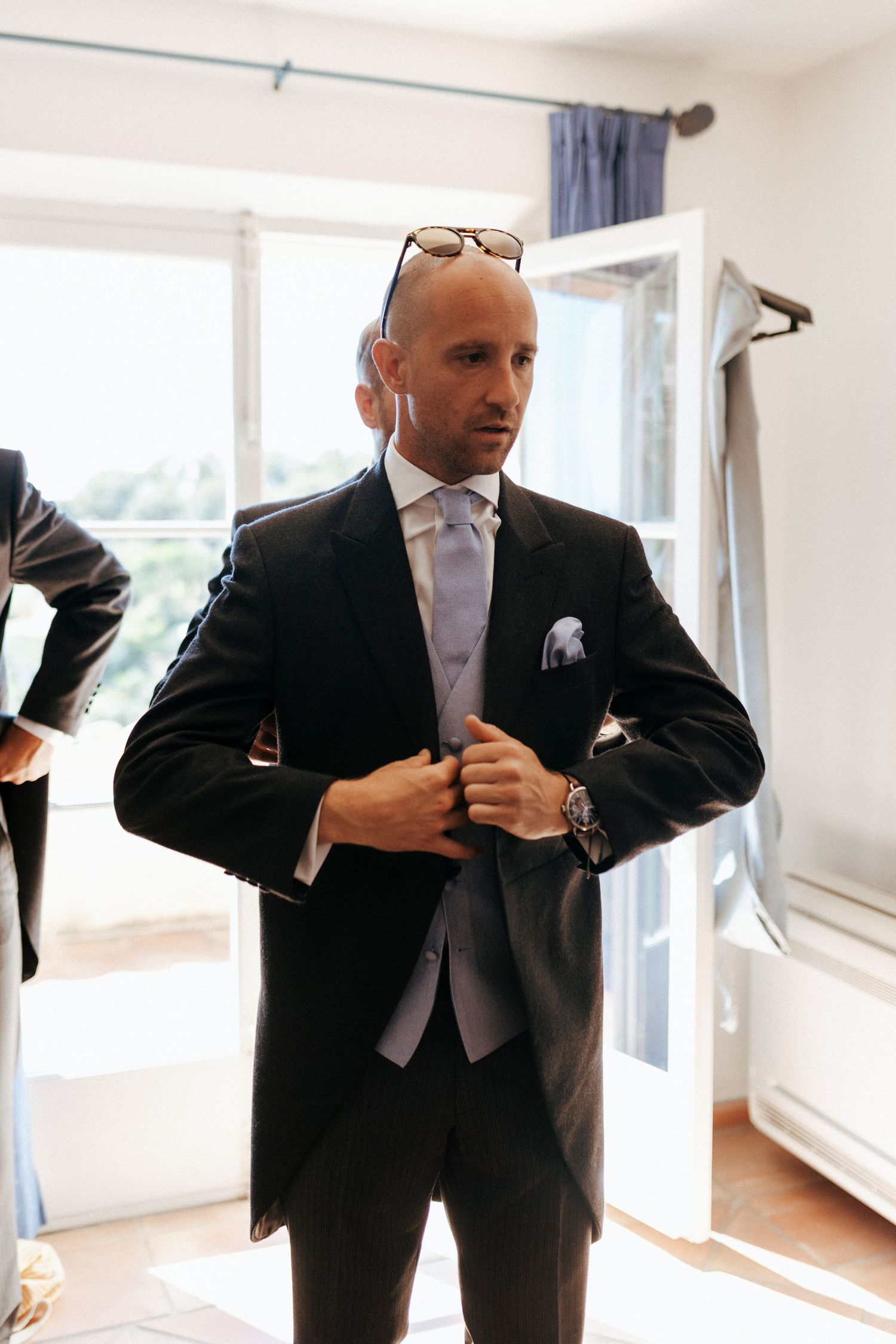 Groom adjusts formal black tuxedo jacket while getting ready in sunlit room.