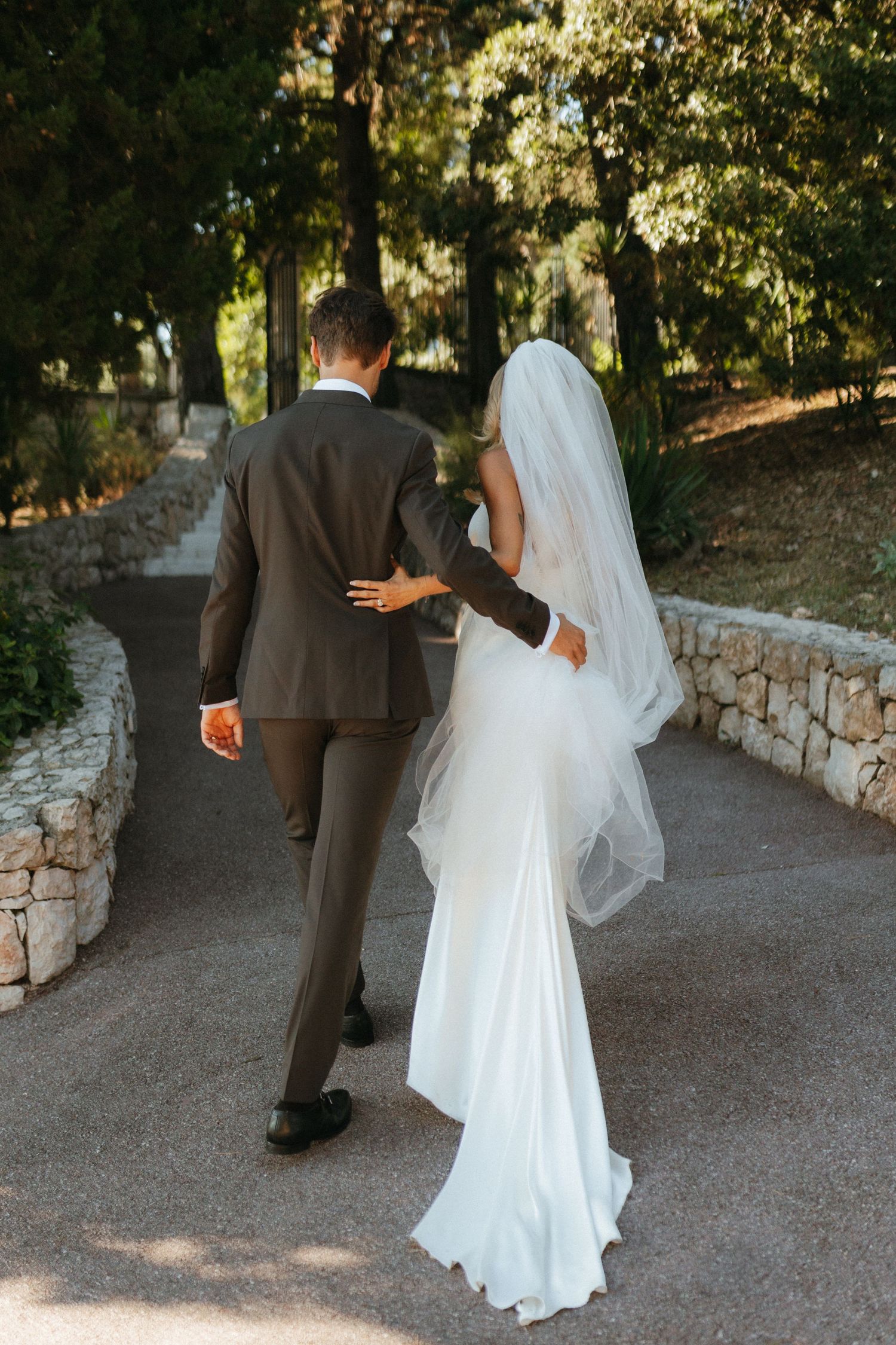 A bride and groom walk together down a stone-lined path through a sunlit garden on their wedding day.