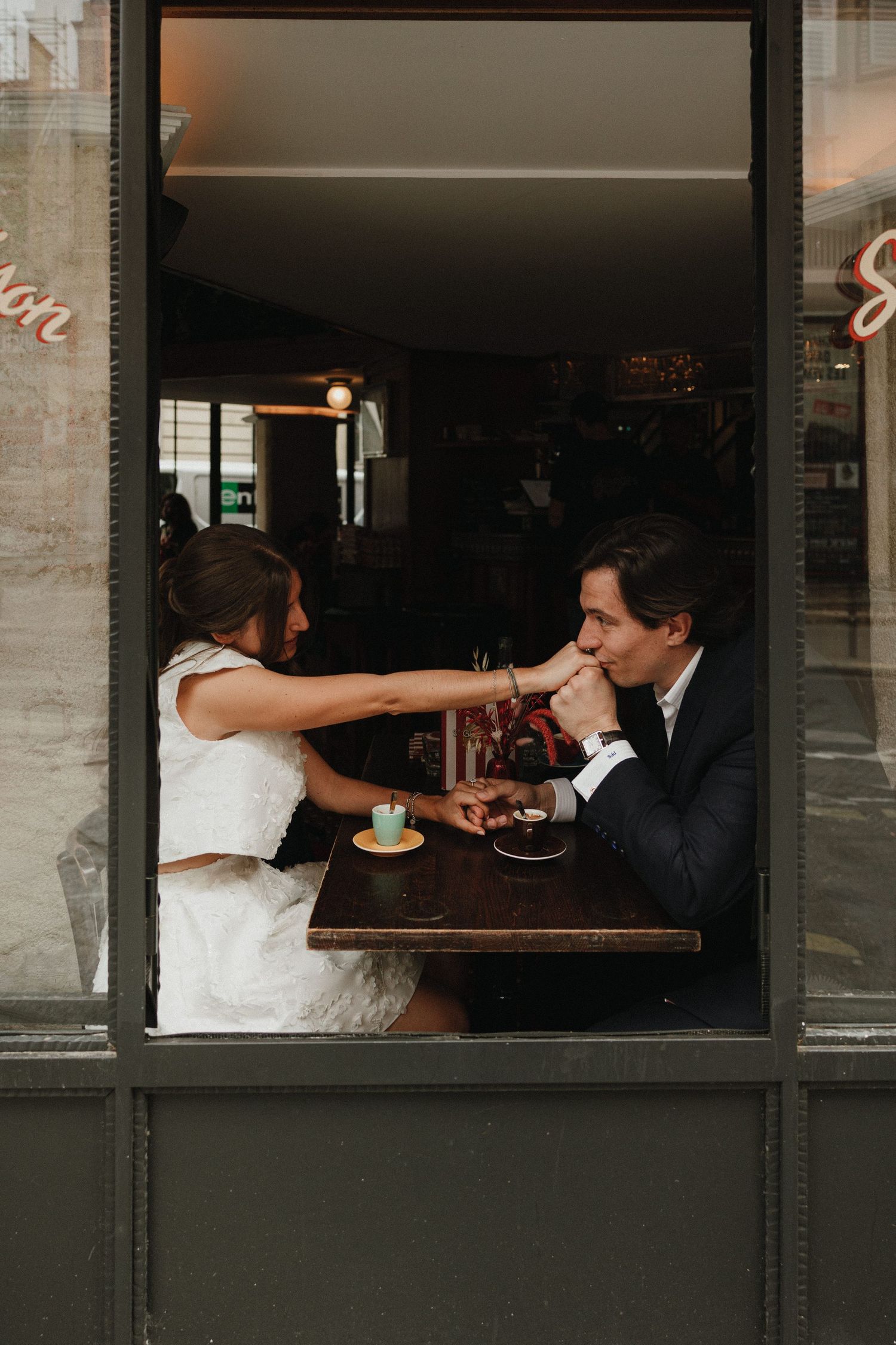 A couple shares a romantic moment at a restaurant table while feeding each other dessert through a window view.