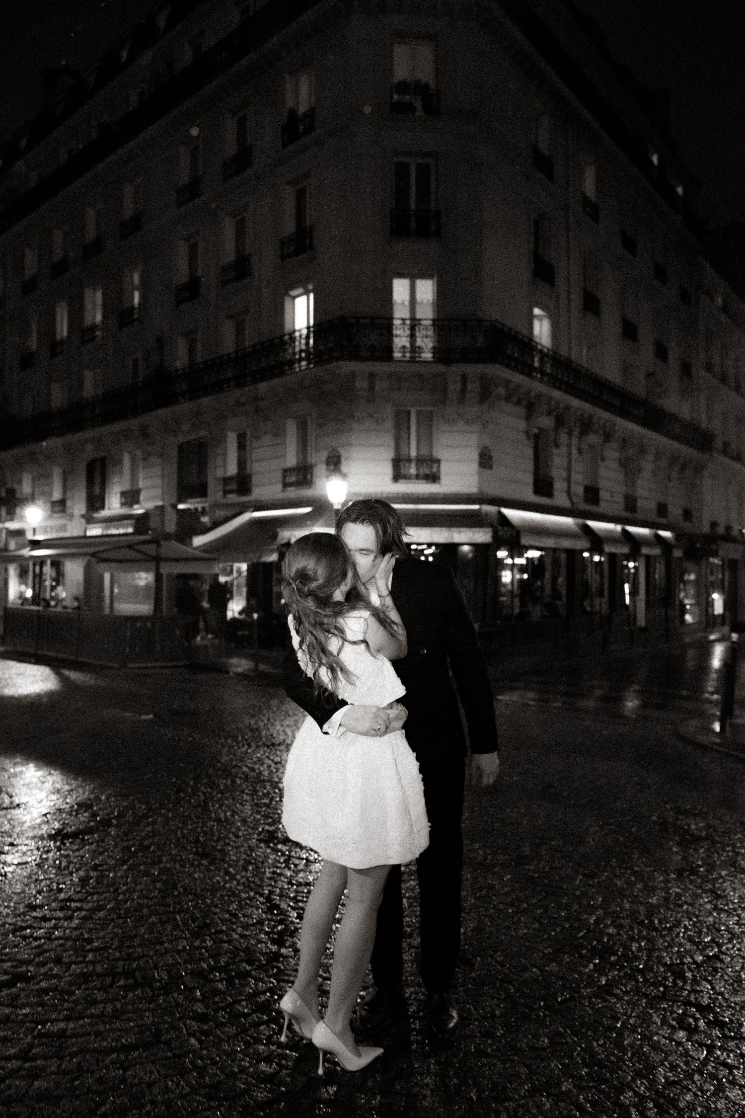 A romantic couple embraces on a rainy cobblestone street at night in Paris with illuminated buildings in the background.