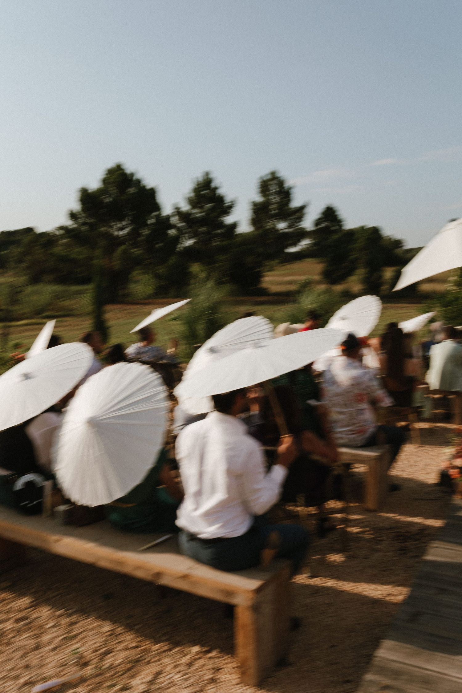 Wedding guests sit on wooden benches holding white paper parasols during an outdoor ceremony.