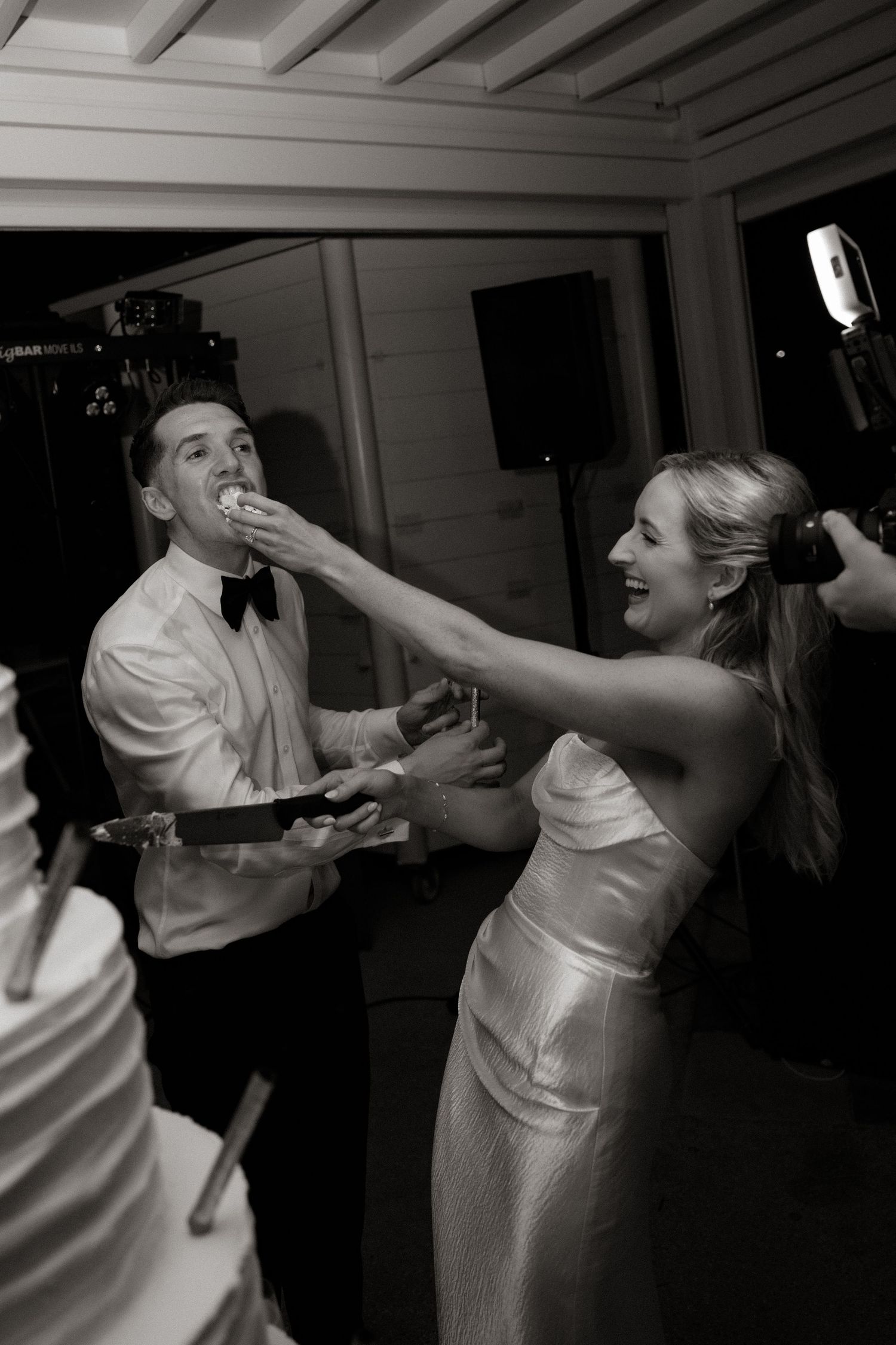 Black and white photo of wedding couple playfully sharing cake during their reception celebration.