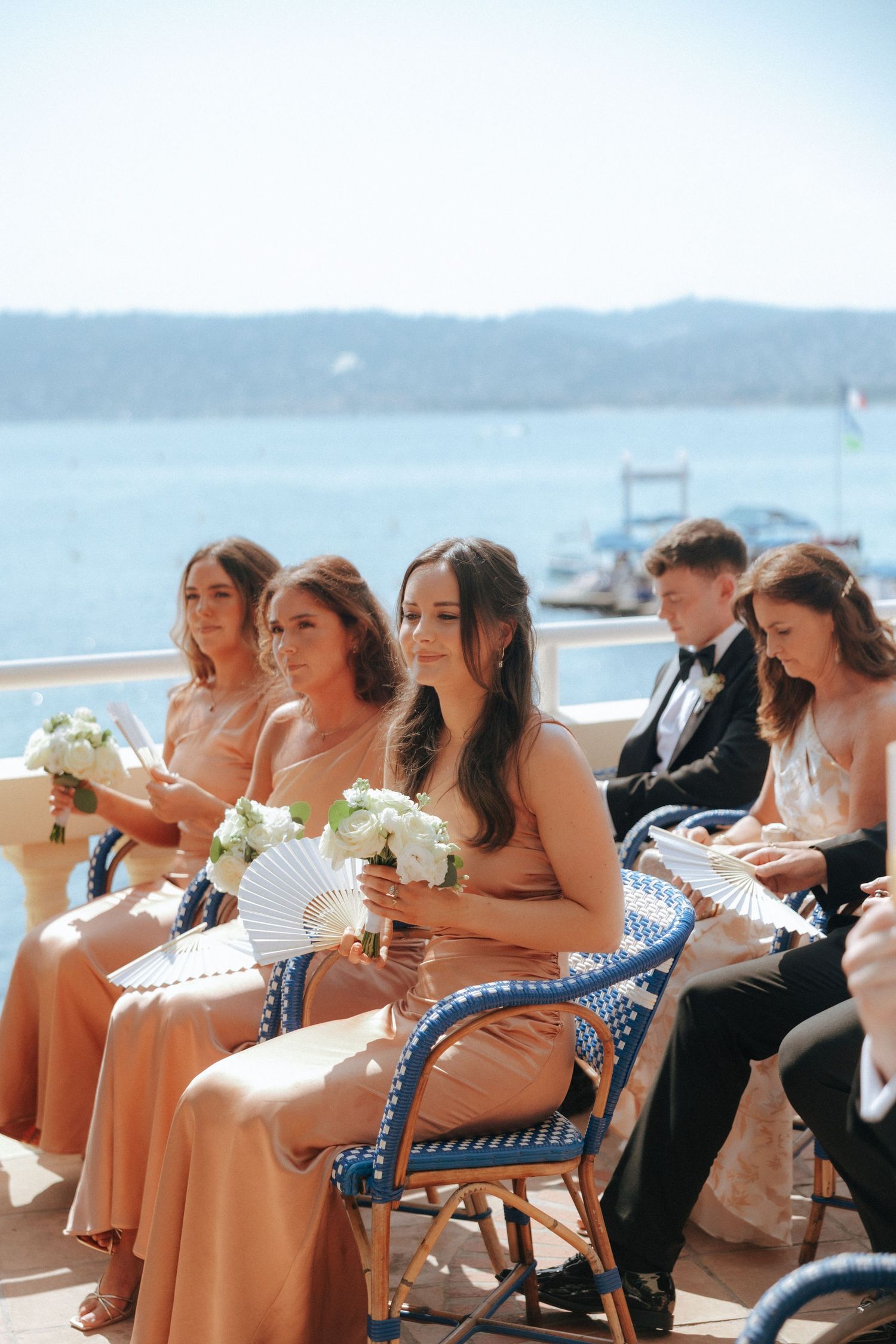 Wedding party seated at outdoor reception with white fans and ocean backdrop.