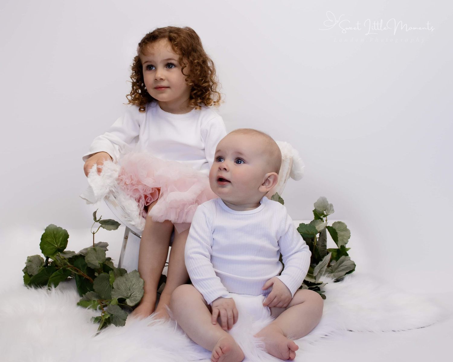 Two children in white outfits posing with greenery on white background.