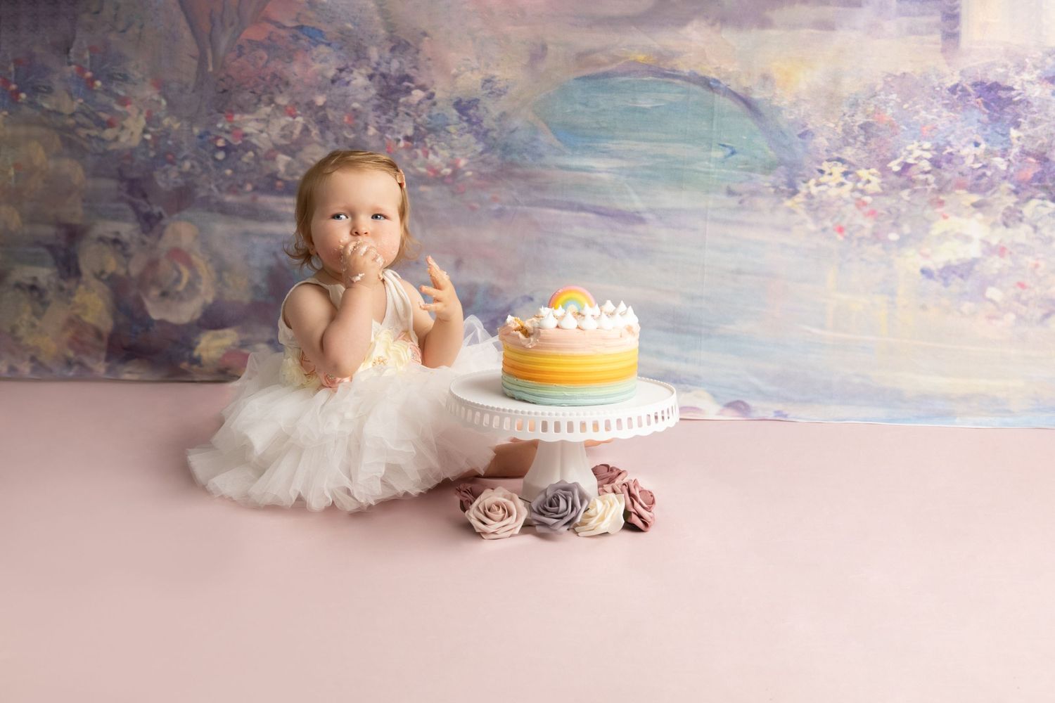 Baby wearing white tutu dress sitting next to birthday cake with floral backdrop.