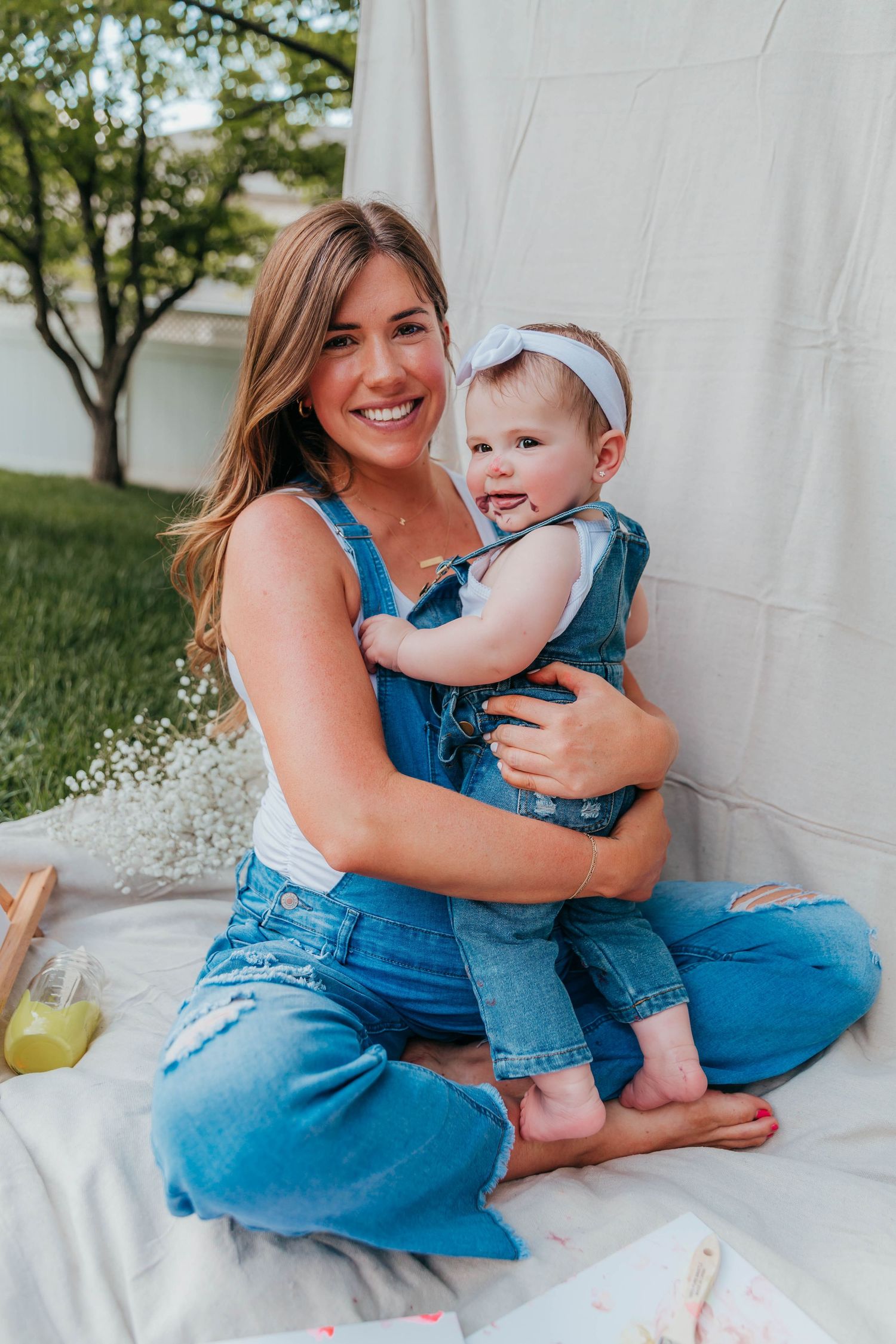 Smiling mom in denim overalls sitting on a blanket in the backyard, holding her toddler who has paint on her face from a Mommy & Me painting