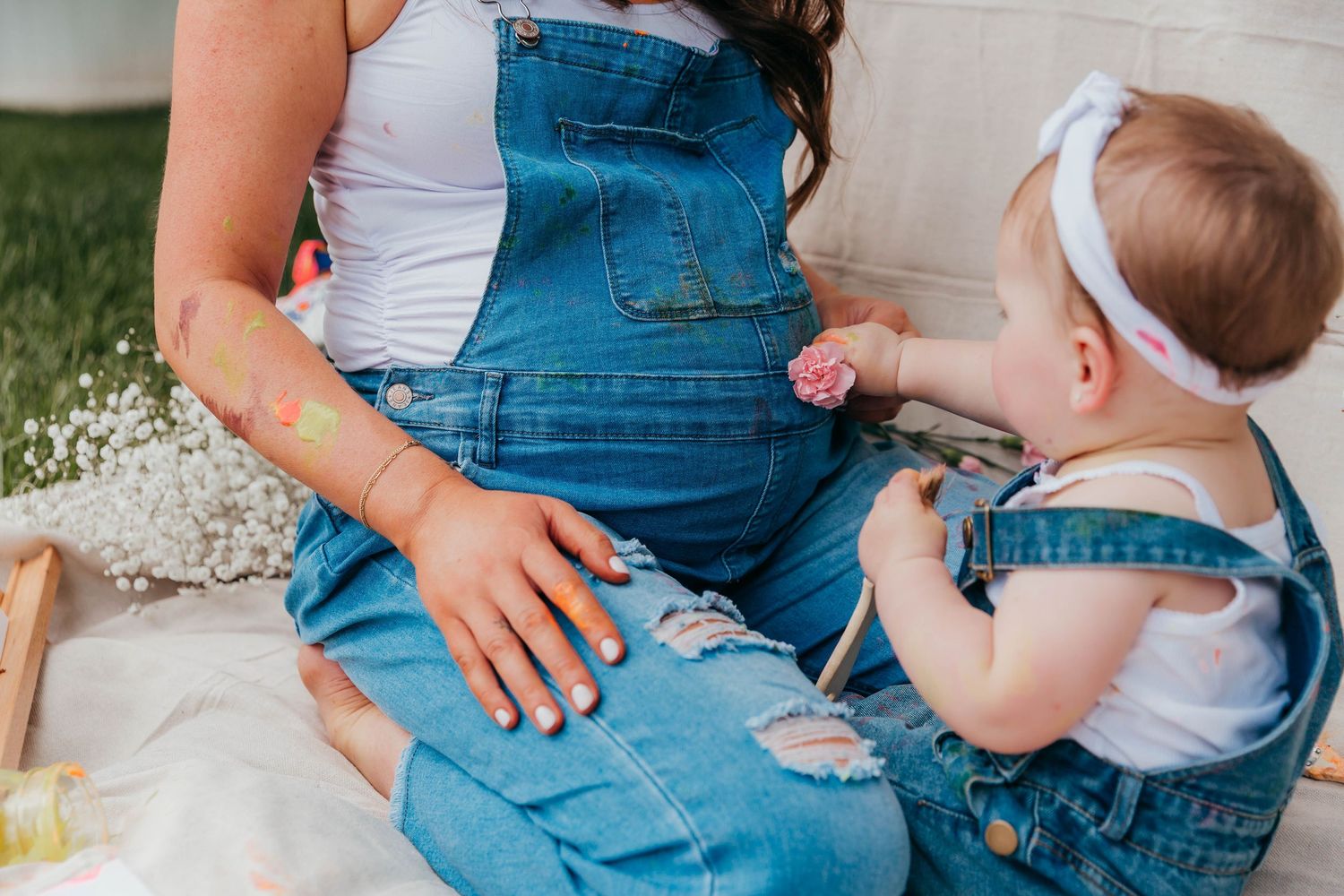 Smiling mom in denim overalls sitting on a blanket in the backyard, holding her toddler who has paint on her face from a Mommy & Me painting
