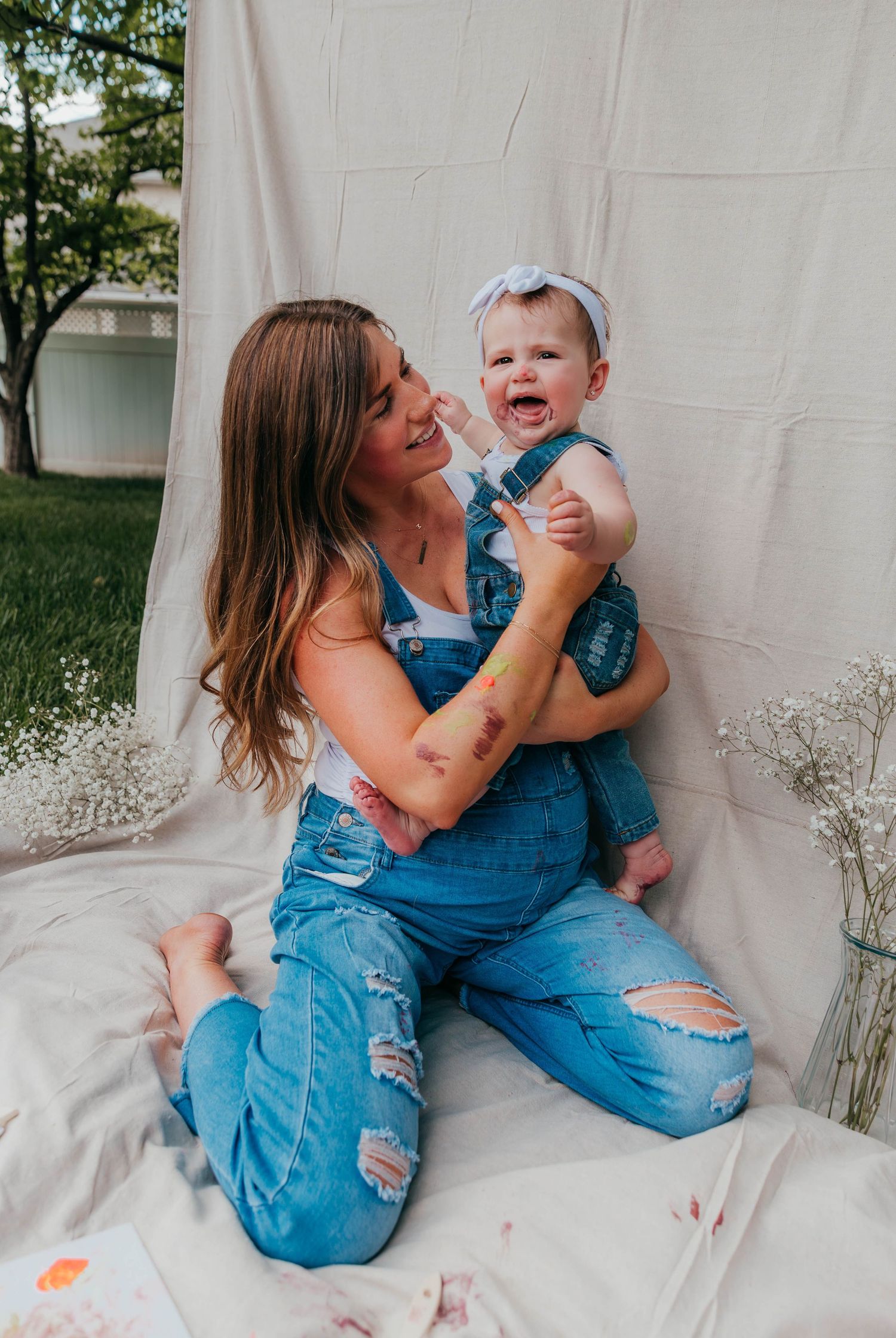 Smiling mom in denim overalls sitting on a blanket in the backyard, holding her toddler who has paint on her face from a Mommy & Me painting