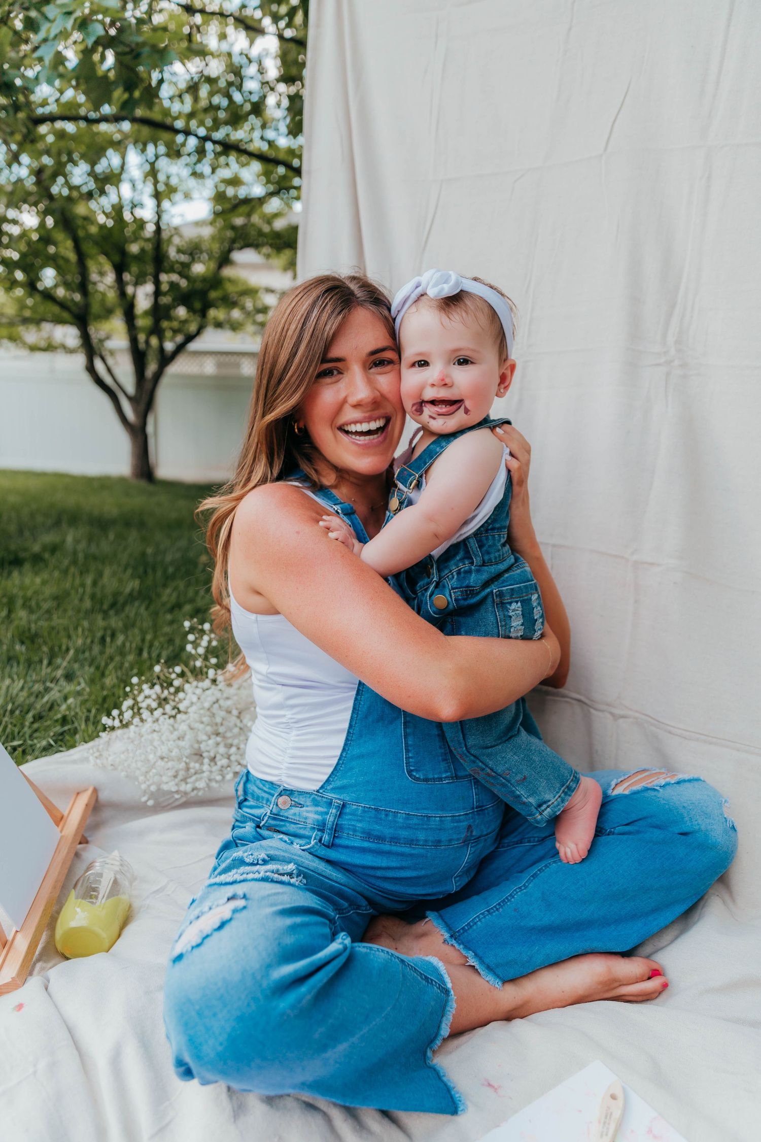 Smiling mom in denim overalls sitting on a blanket in the backyard, holding her toddler who has paint on her face from a Mommy & Me painting