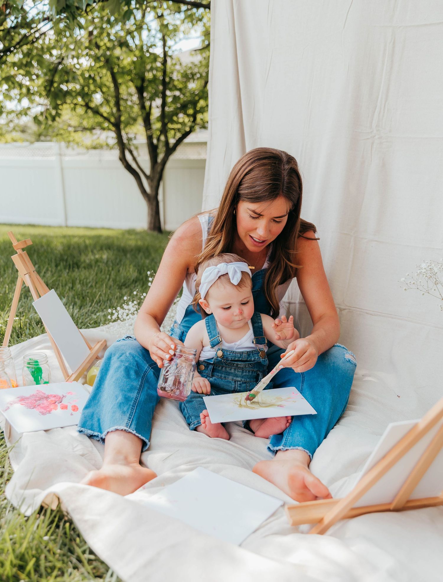 Smiling mom in denim overalls sitting on a blanket in the backyard, holding her toddler who has paint on her face from a Mommy & Me painting