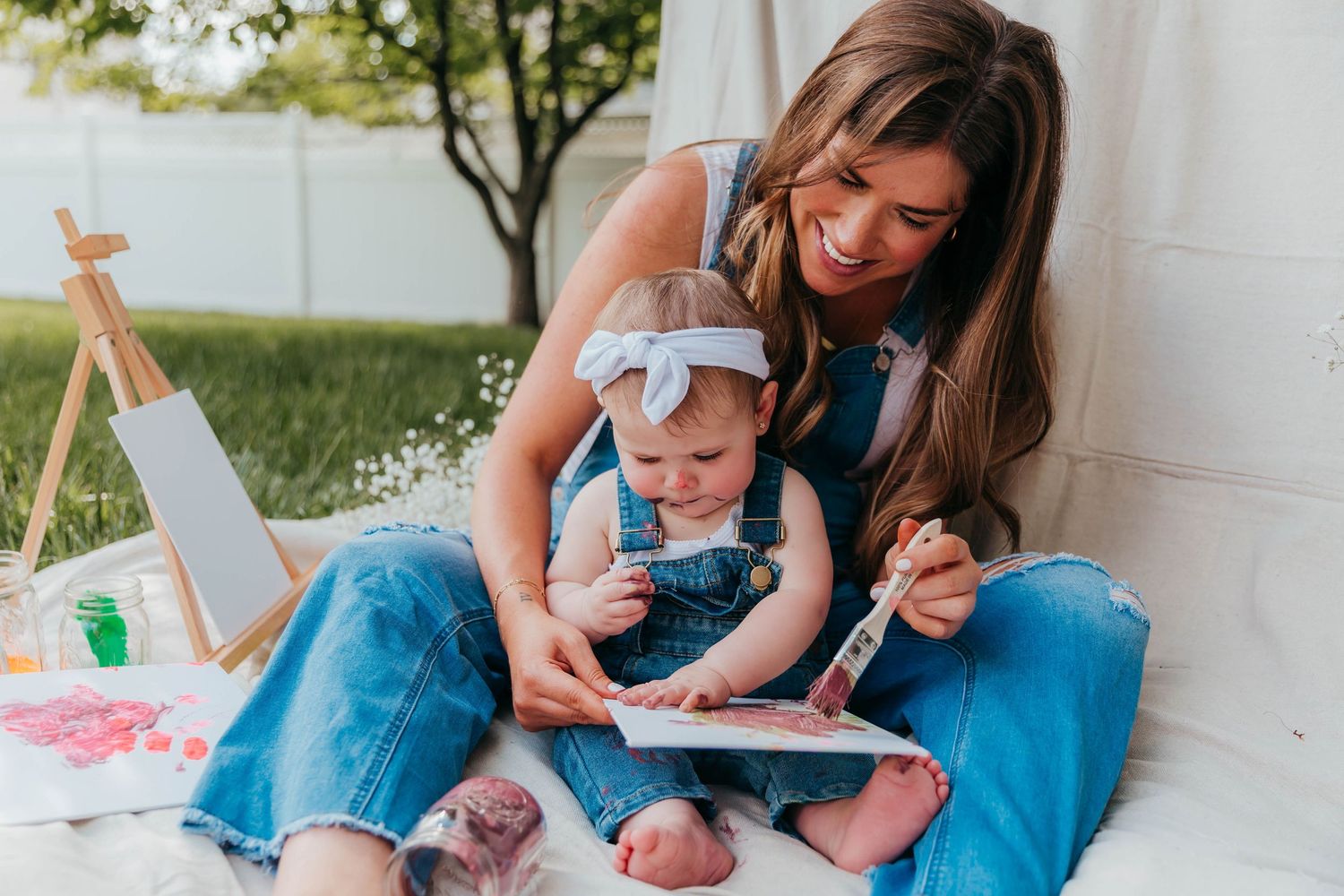 Smiling mom in denim overalls sitting on a blanket in the backyard, holding her toddler who has paint on her face from a Mommy & Me painting