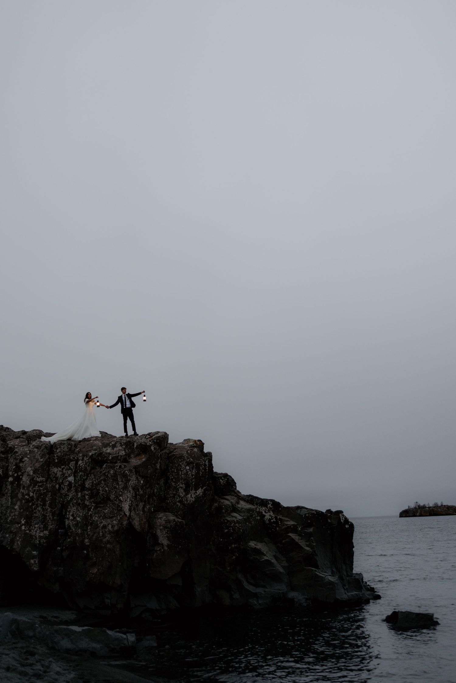 Silhouetted couple stands dramatically on a rocky cliff overlooking misty ocean waters at dusk.