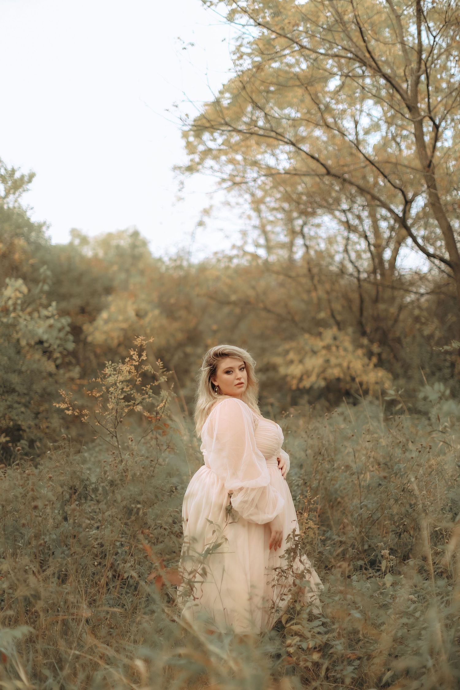A figure stands in a dreamy autumn field wearing a flowing pink dress among tall grass and golden trees.