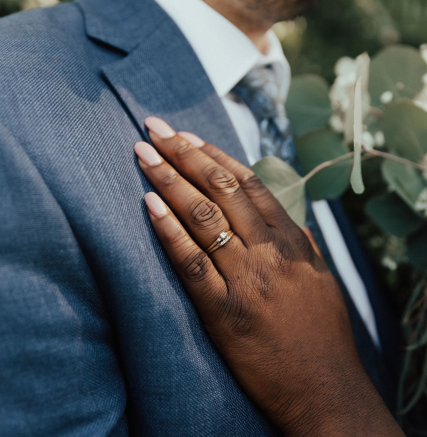 Hand wearing wedding rings gently touching a blue suit jacket sleeve in a romantic close-up shot.