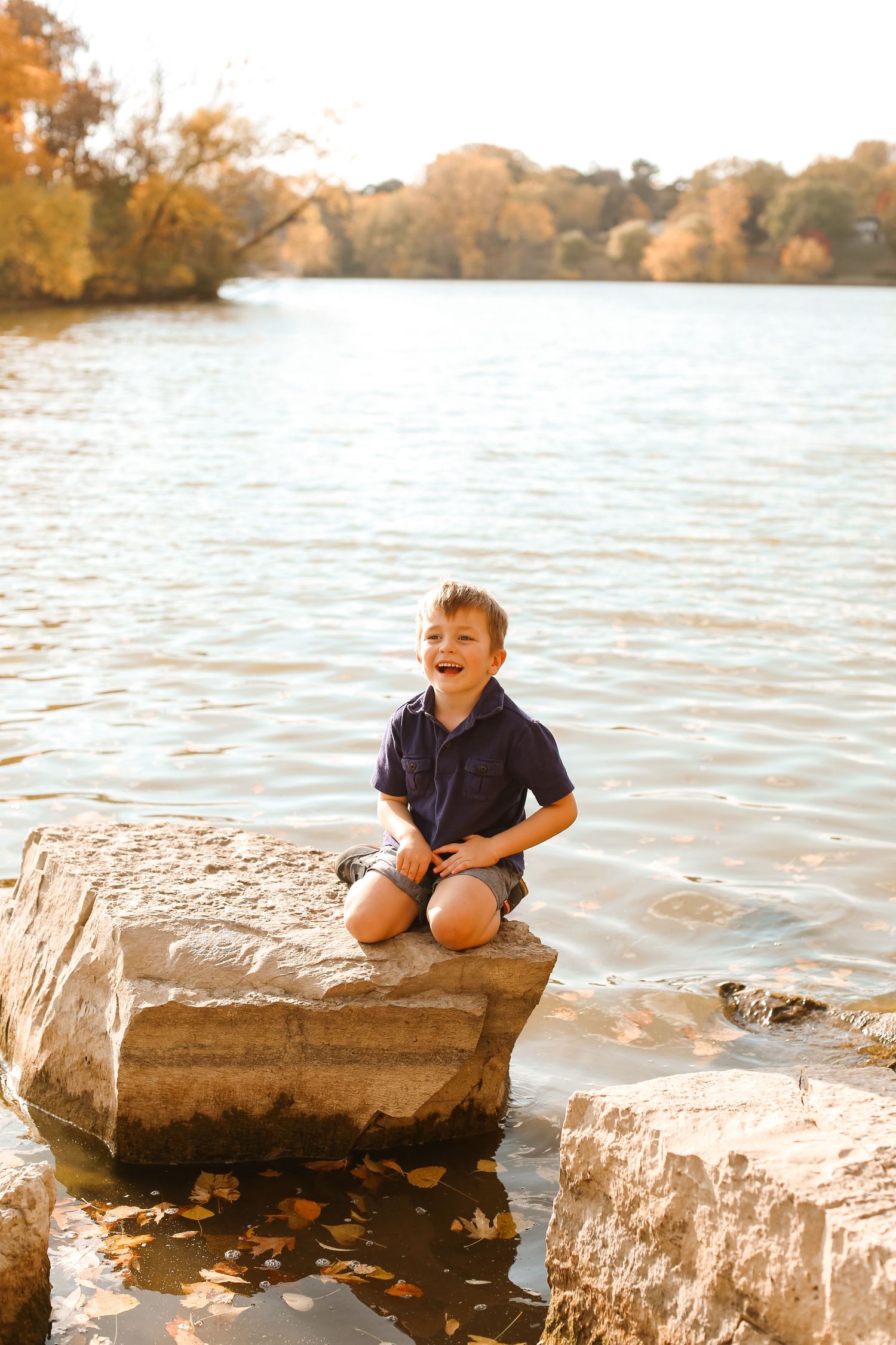 Young boy sits joyfully on rock by lake during golden sunset.