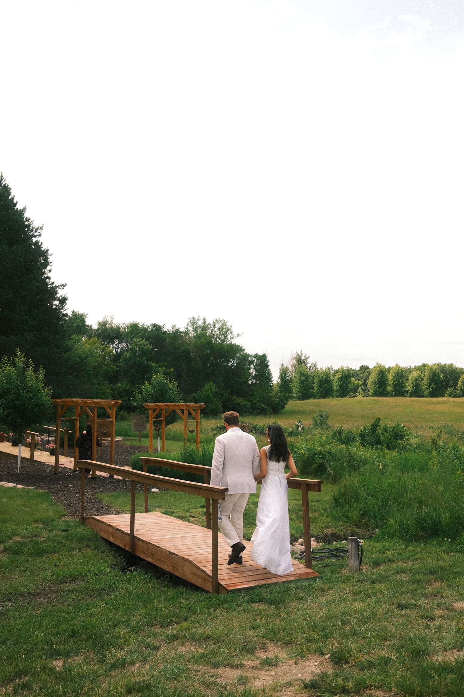 A couple stands on a wooden bridge walkway surrounded by green meadows and trees on a summer day.