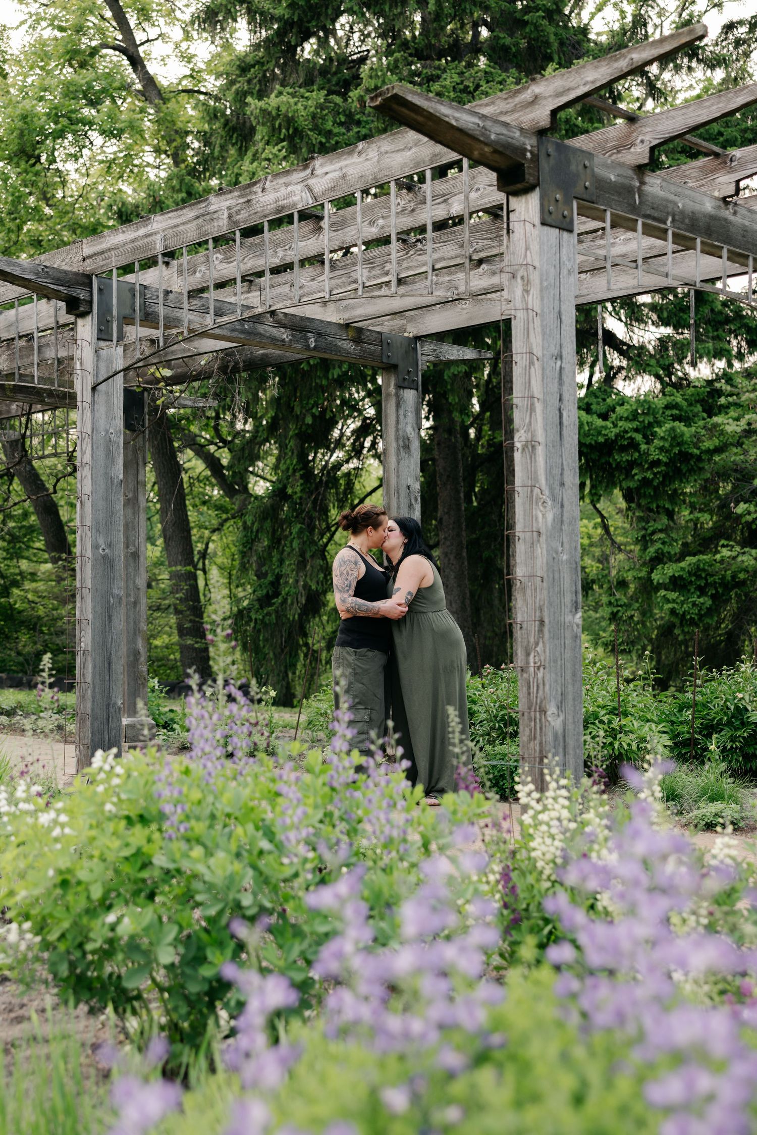 A romantic moment under a wooden pergola surrounded by blooming purple wildflowers in a lush garden setting.