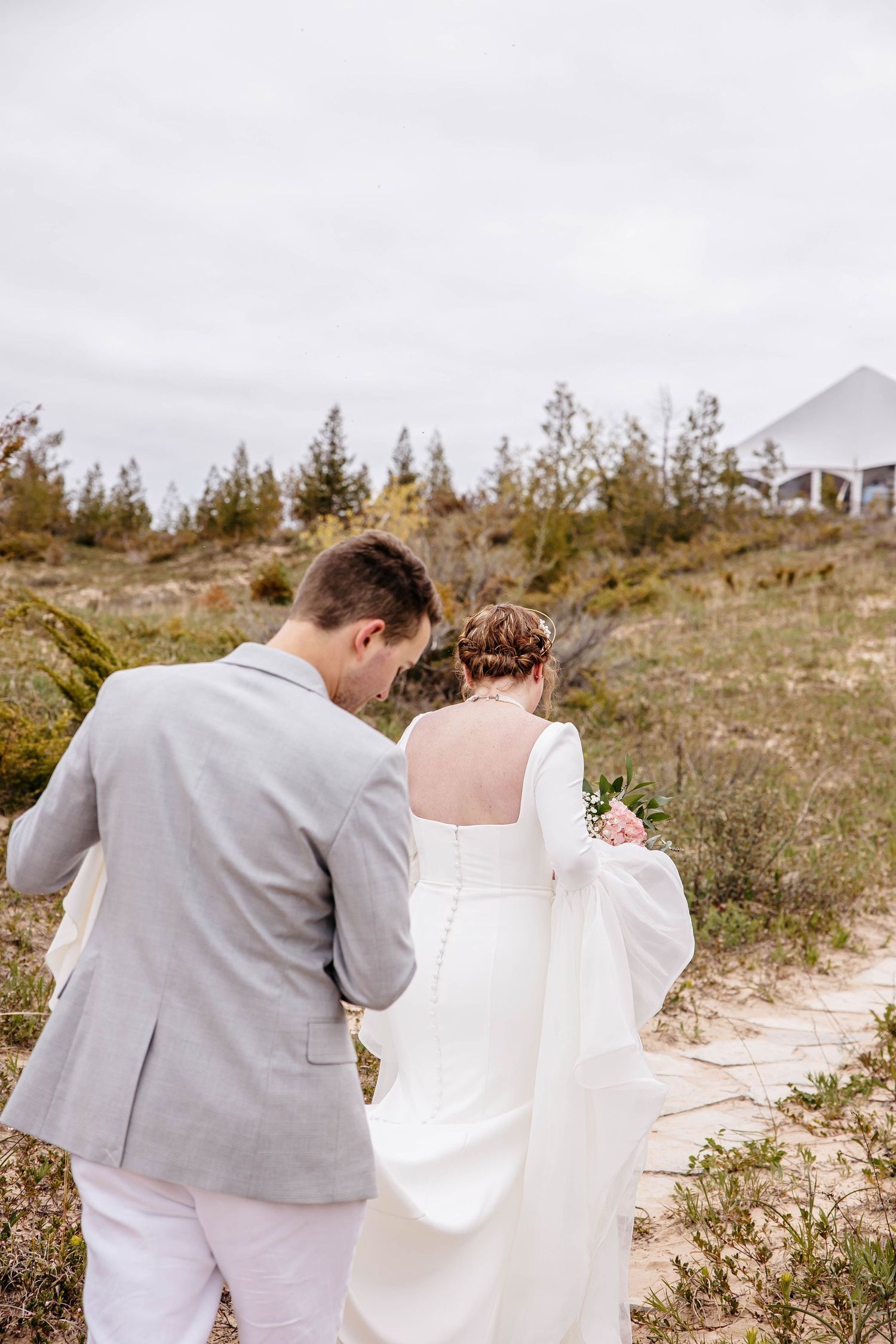 Couple walking along a sandy beach path with the person in gray holding up the trailing white dress.
