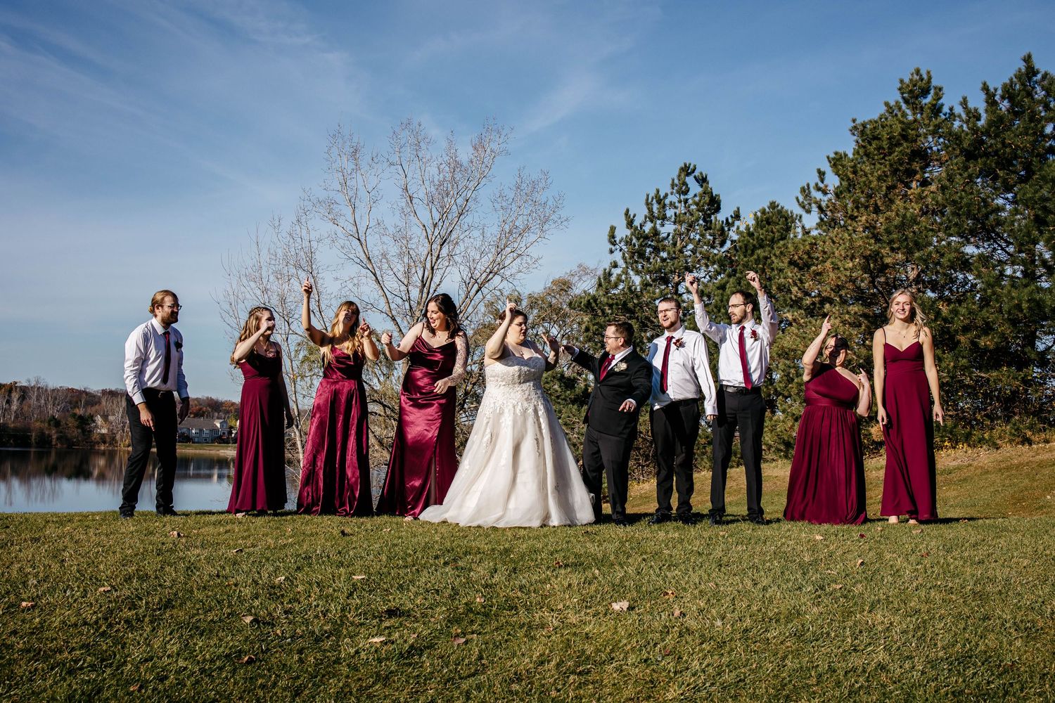 Wedding party standing in a line outdoors against blue sky with bridesmaids in burgundy dresses and groomsmen in suits.