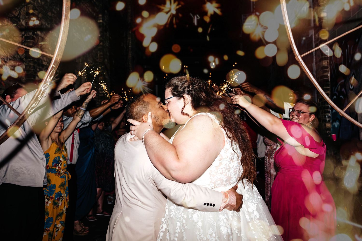 A couple stands under romantic lighting and bubbles while embracing during a nighttime wedding celebration.