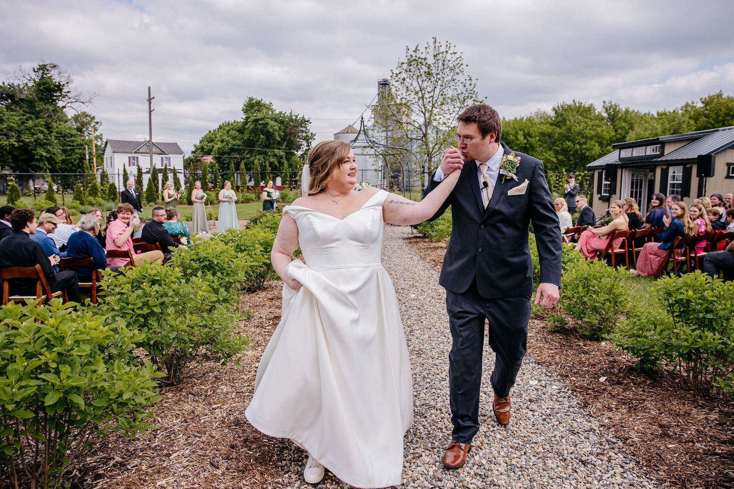 Two people walk joyfully down a gravel path surrounded by garden plants and guests at an outdoor event.